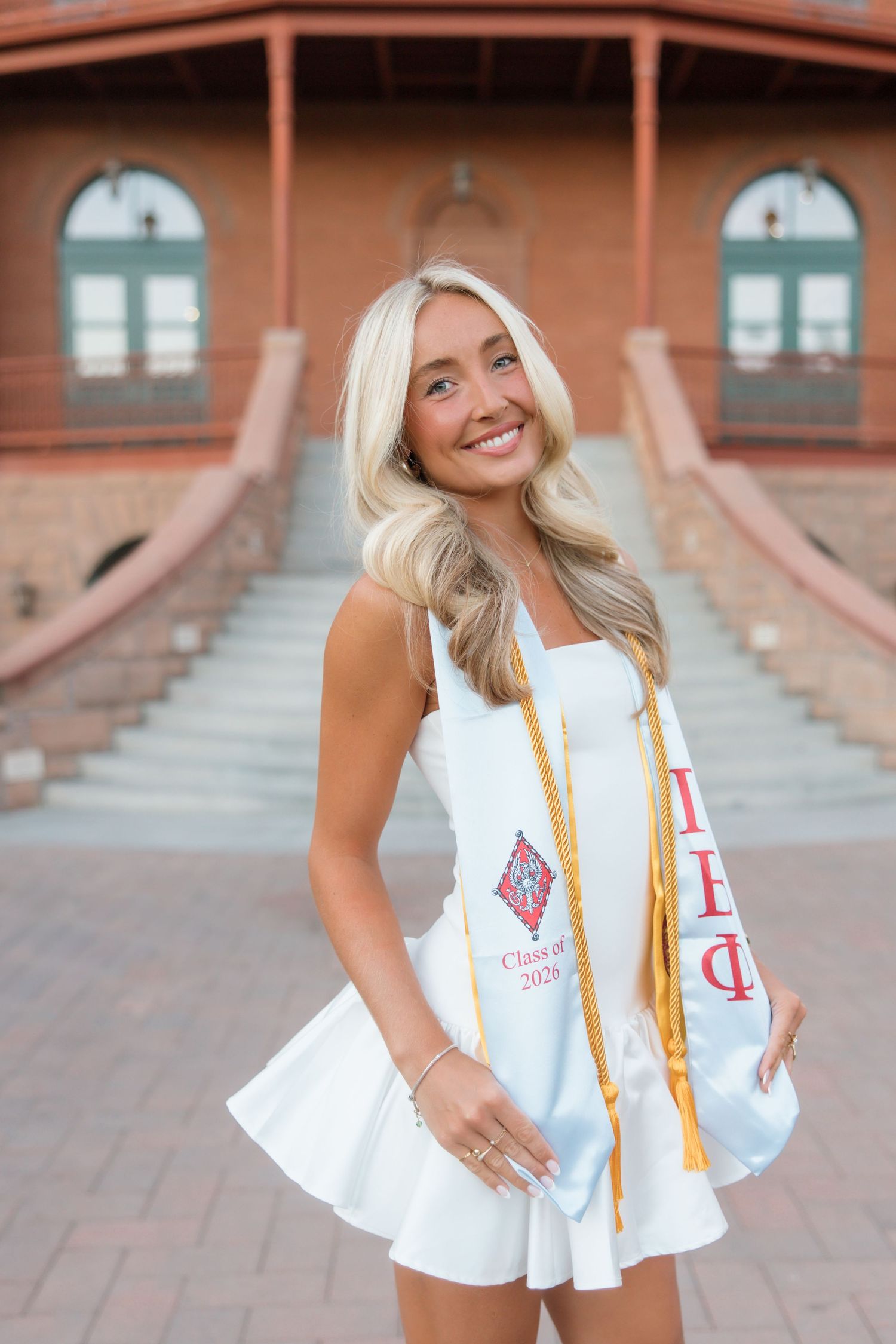 Smiling blonde woman in white dress wearing graduation stole and gold tassel, posing outdoors at Class of 2024 ceremony.
