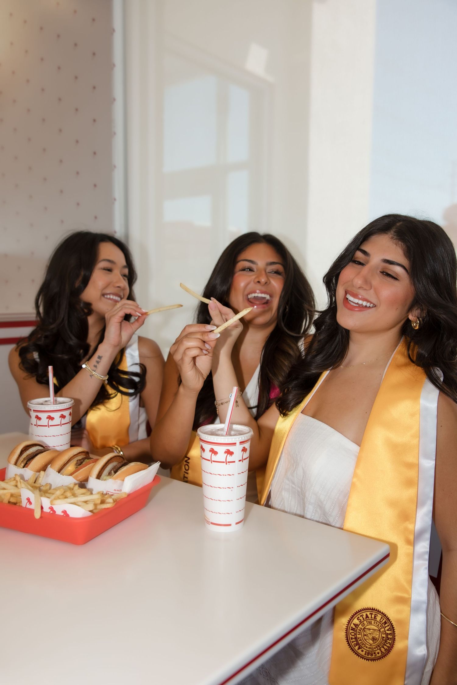 Three smiling graduates in white dresses and gold stoles enjoy fries and drinks inside an In-N-Out Burger.