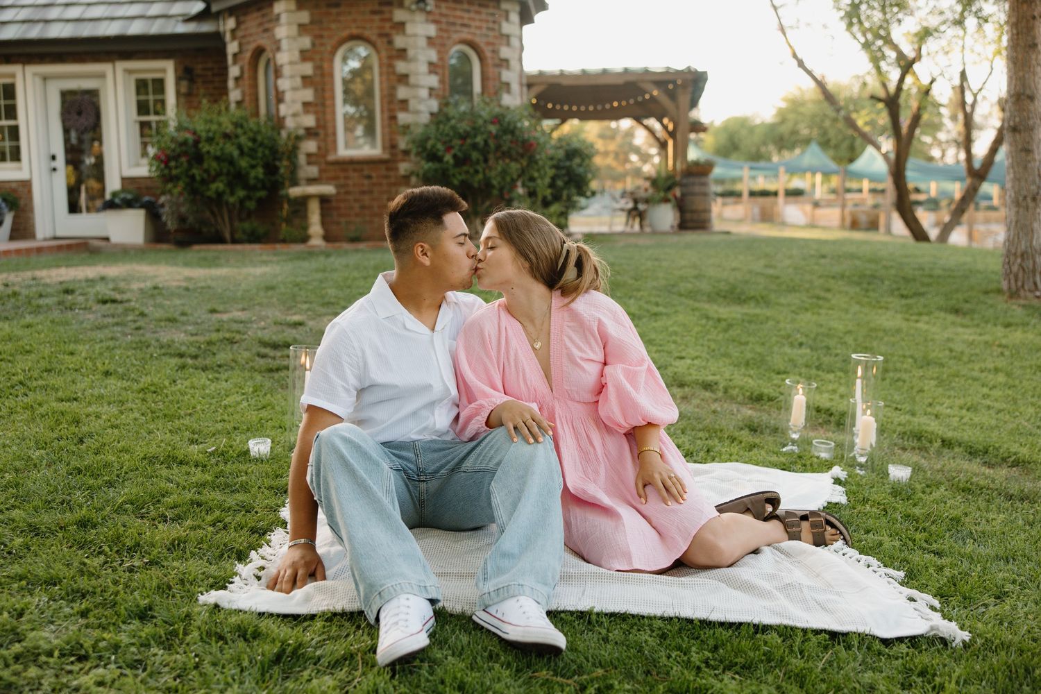 Couple sharing a romantic kiss during a picnic on a green lawn outside a brick building at sunset.