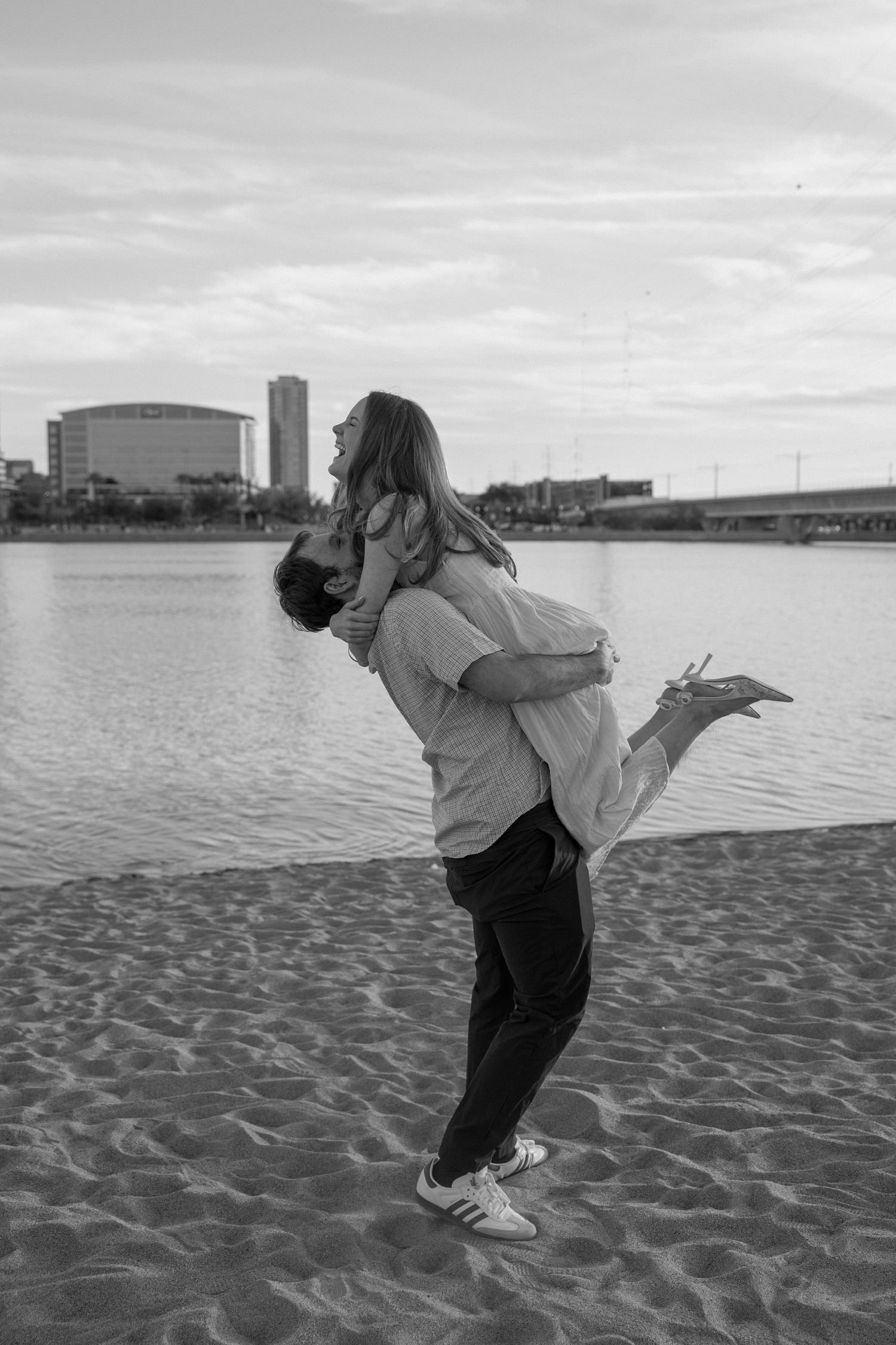Black and white photo of a couple on a sandy beach, man lifting woman as she laughs joyfully.