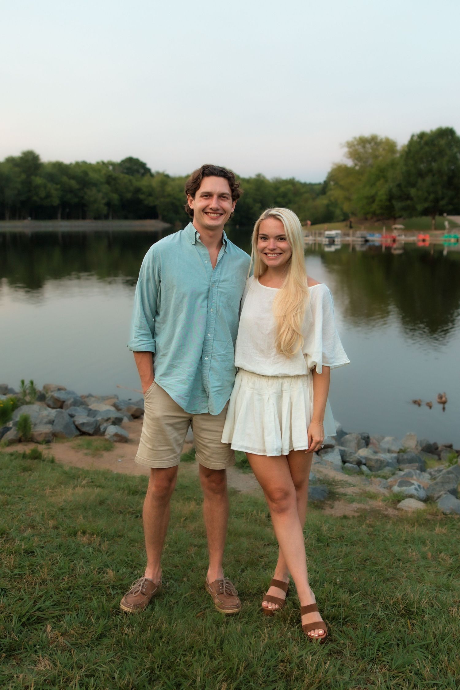 Smiling young couple standing together by a rocky lakeside at sunset in a park.