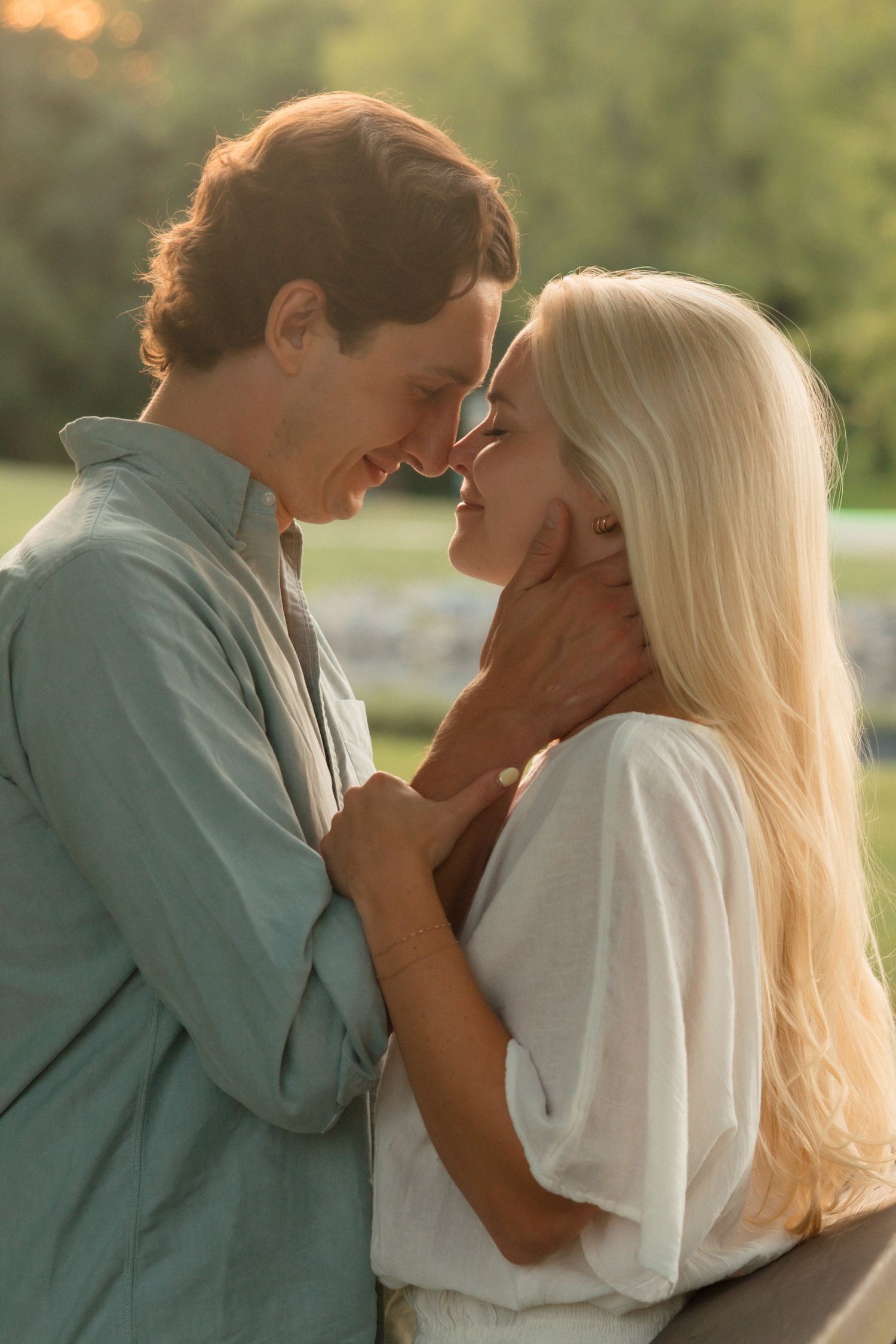 A romantic couple leans in for a kiss at golden hour, touching foreheads in a lush green park setting.