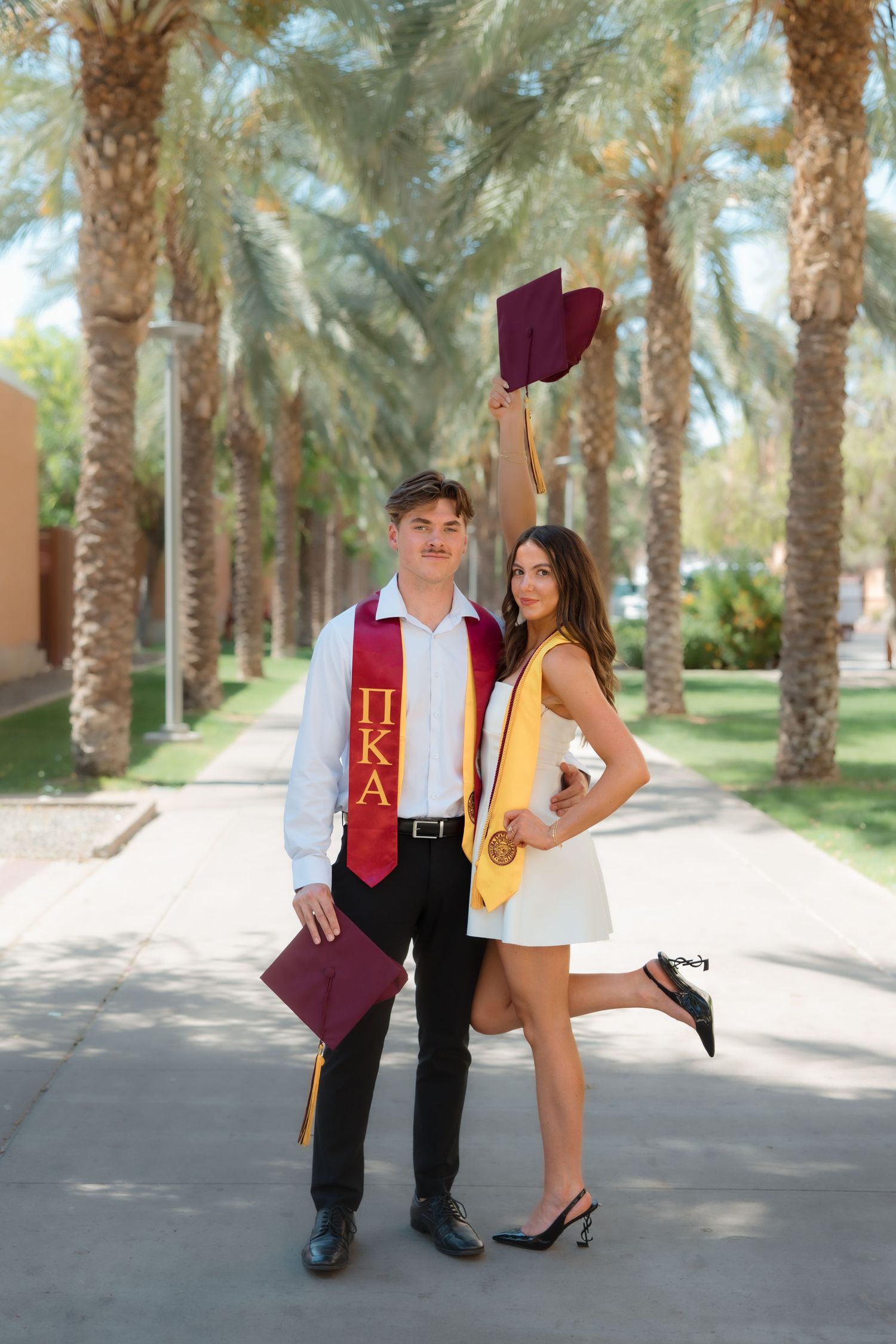 Graduating couple posing on palm tree walkway, man raising maroon cap in celebration.