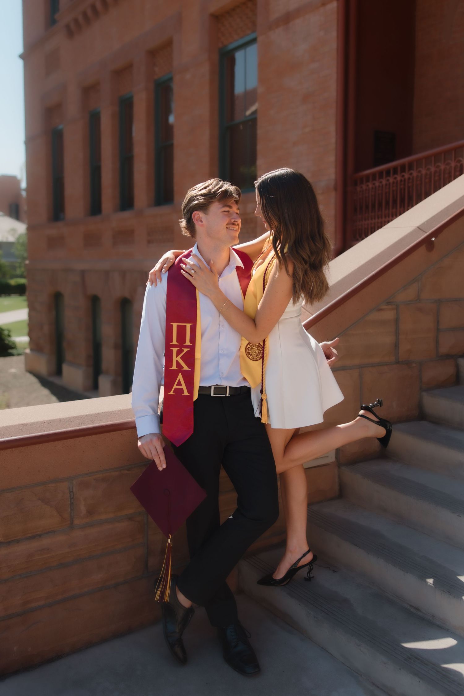 Couple in graduation stoles share romantic moment leaning against brick wall on college campus.