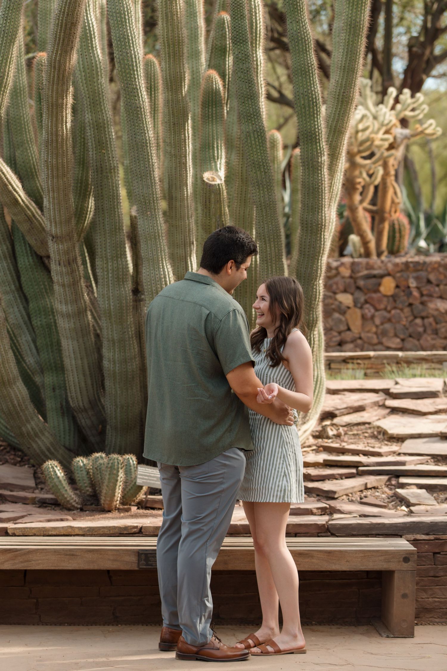 Couple holding hands and smiling at each other in front of tall cacti at a desert botanical garden.