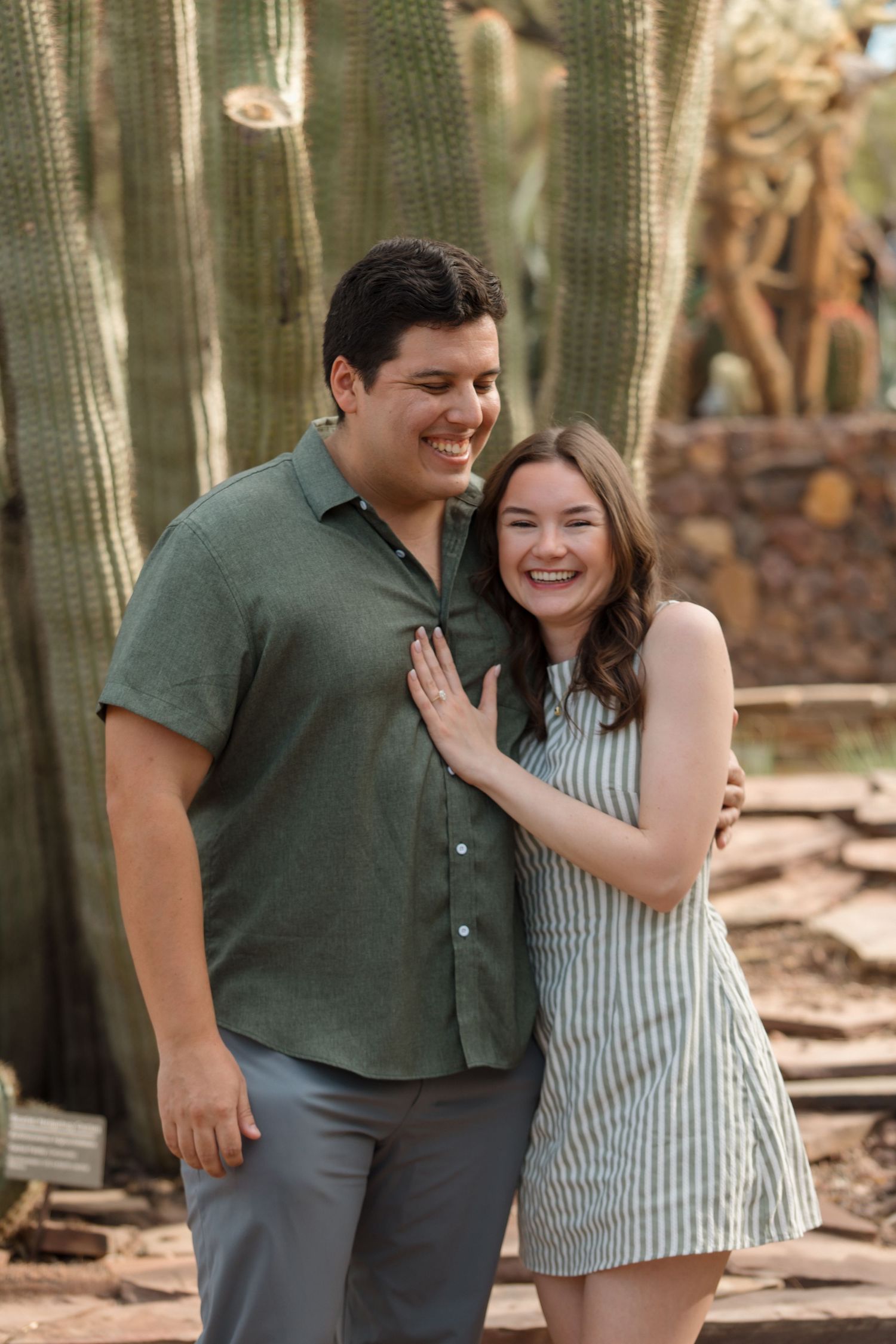 Smiling couple embracing in front of tall cacti, woman in striped dress and man in green shirt.