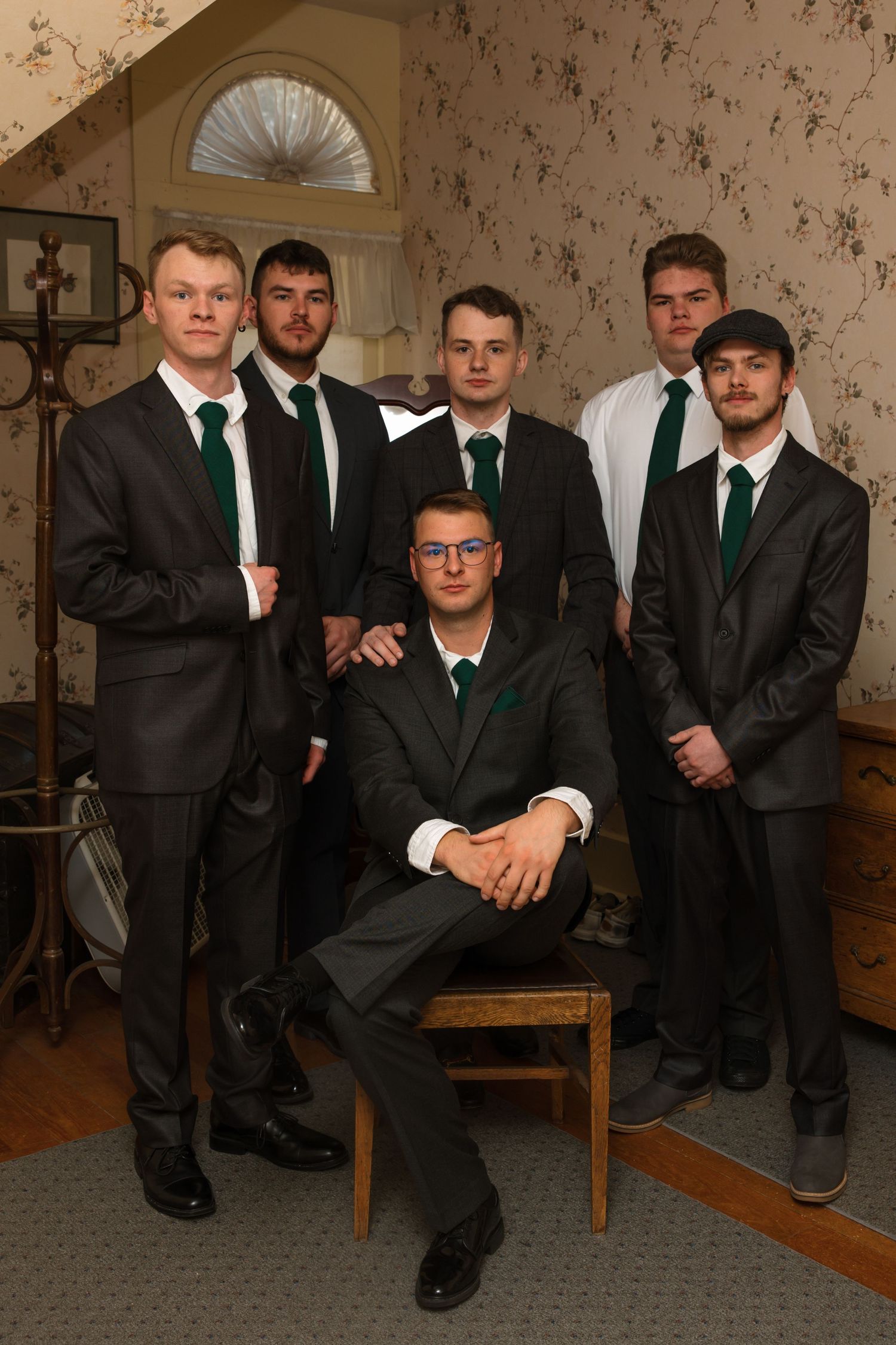 Six groomsmen in dark suits with green ties pose formally in a floral wallpapered room for a wedding photo.
