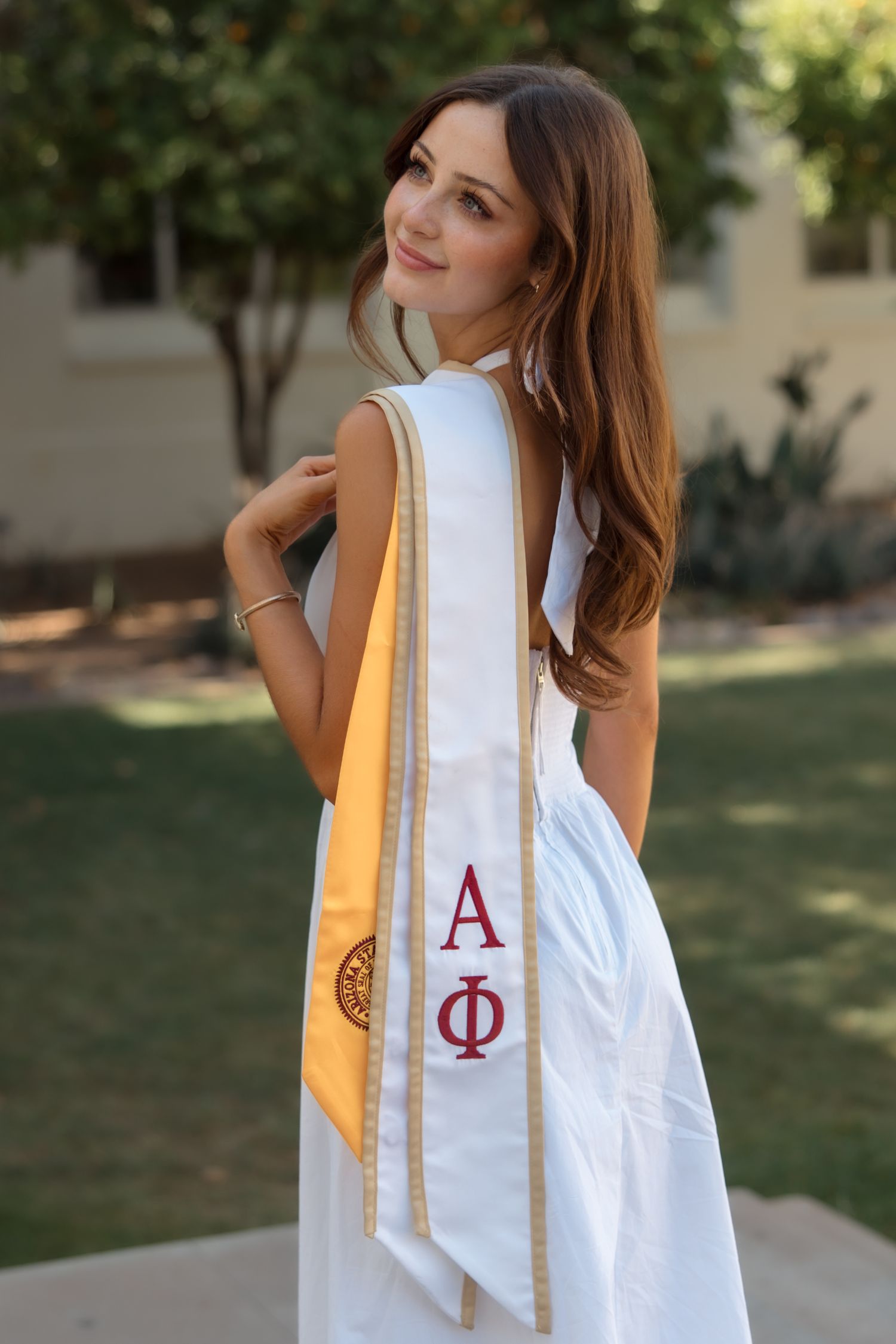 Young woman in white graduation gown wearing Alpha Phi sorority stole, smiling outdoors on a sunny day.