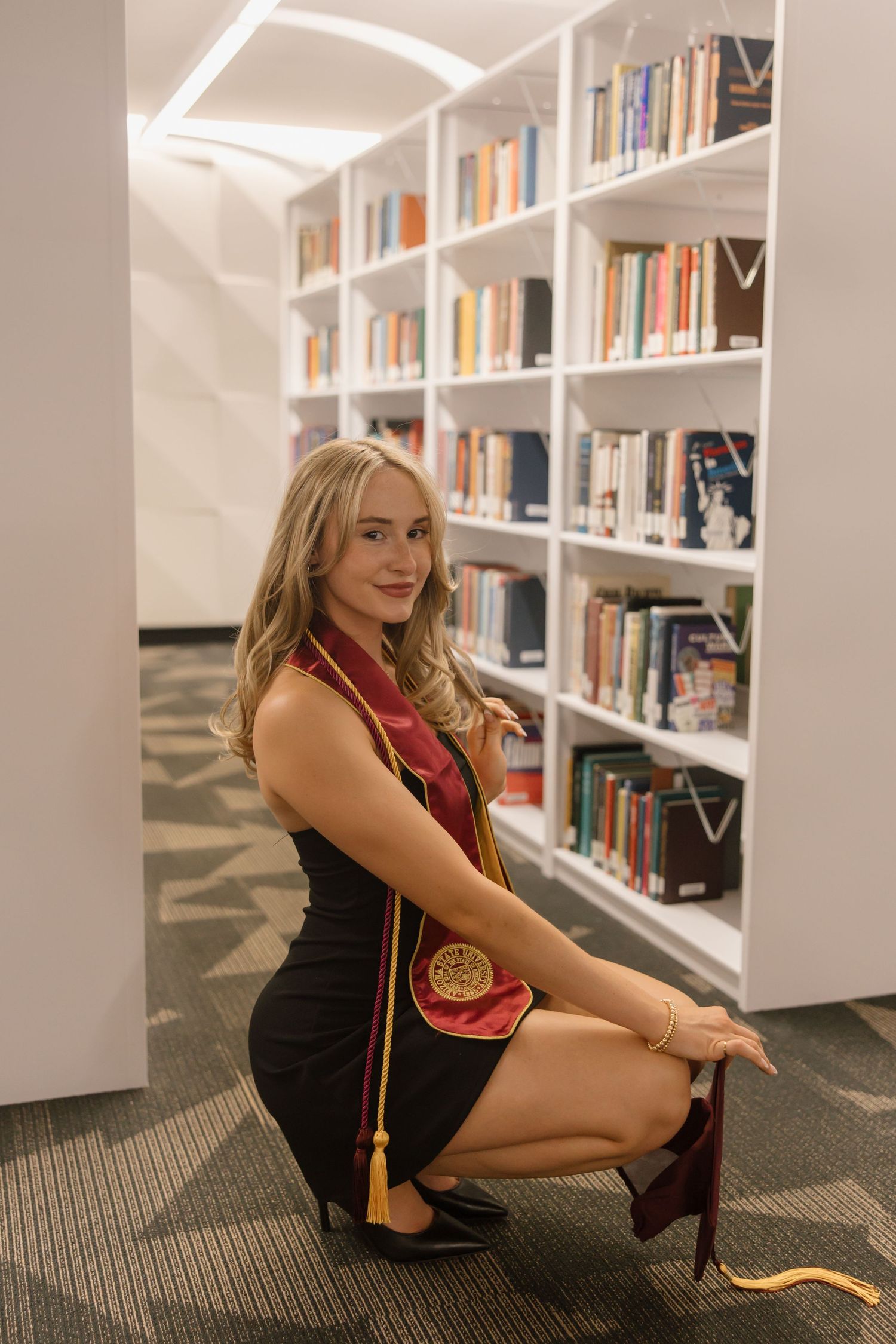 Blonde woman in black dress with maroon graduation stole poses in a modern library for graduation photos.