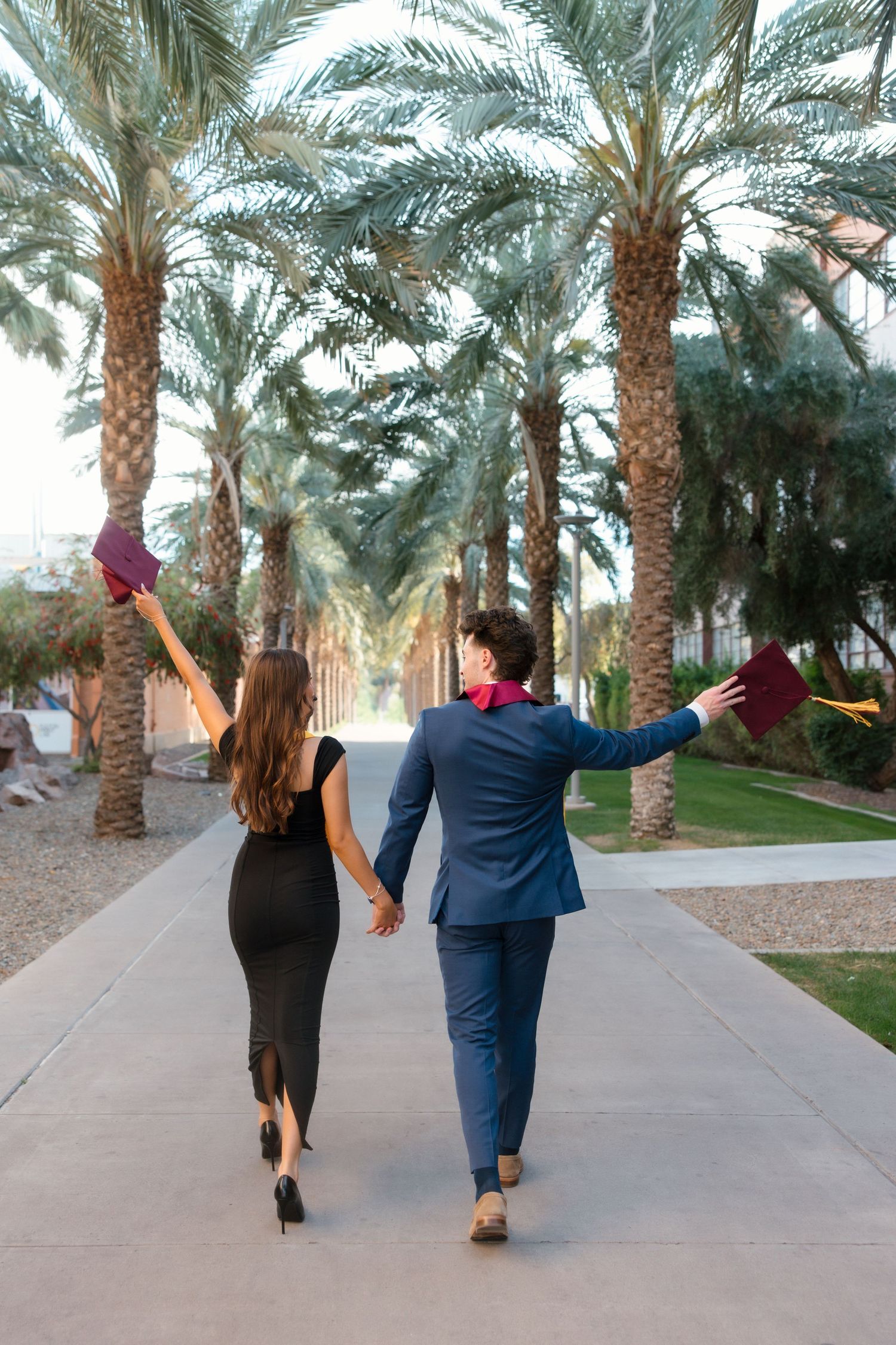 Two graduates in formal attire walk hand-in-hand along a palm tree-lined path, raising their mortarboards in celebration.