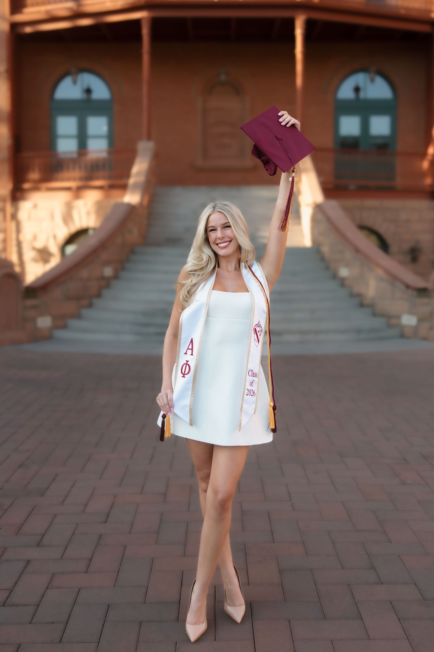 Smiling blonde graduate in white dress holds maroon cap aloft, wearing Alpha Phi sash on brick campus steps.