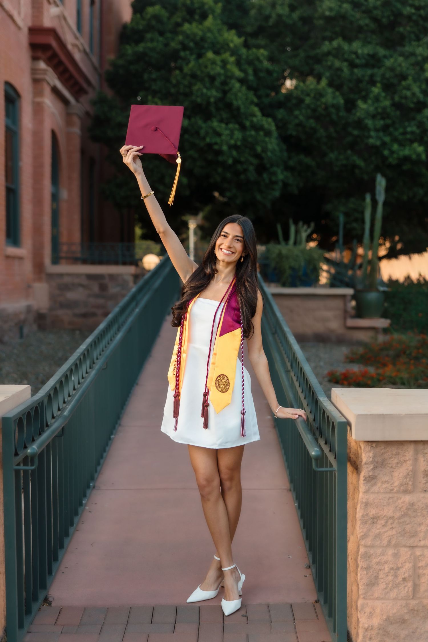 Young woman in white dress holds maroon graduation cap aloft on bridge, wearing ASU sash and stole.