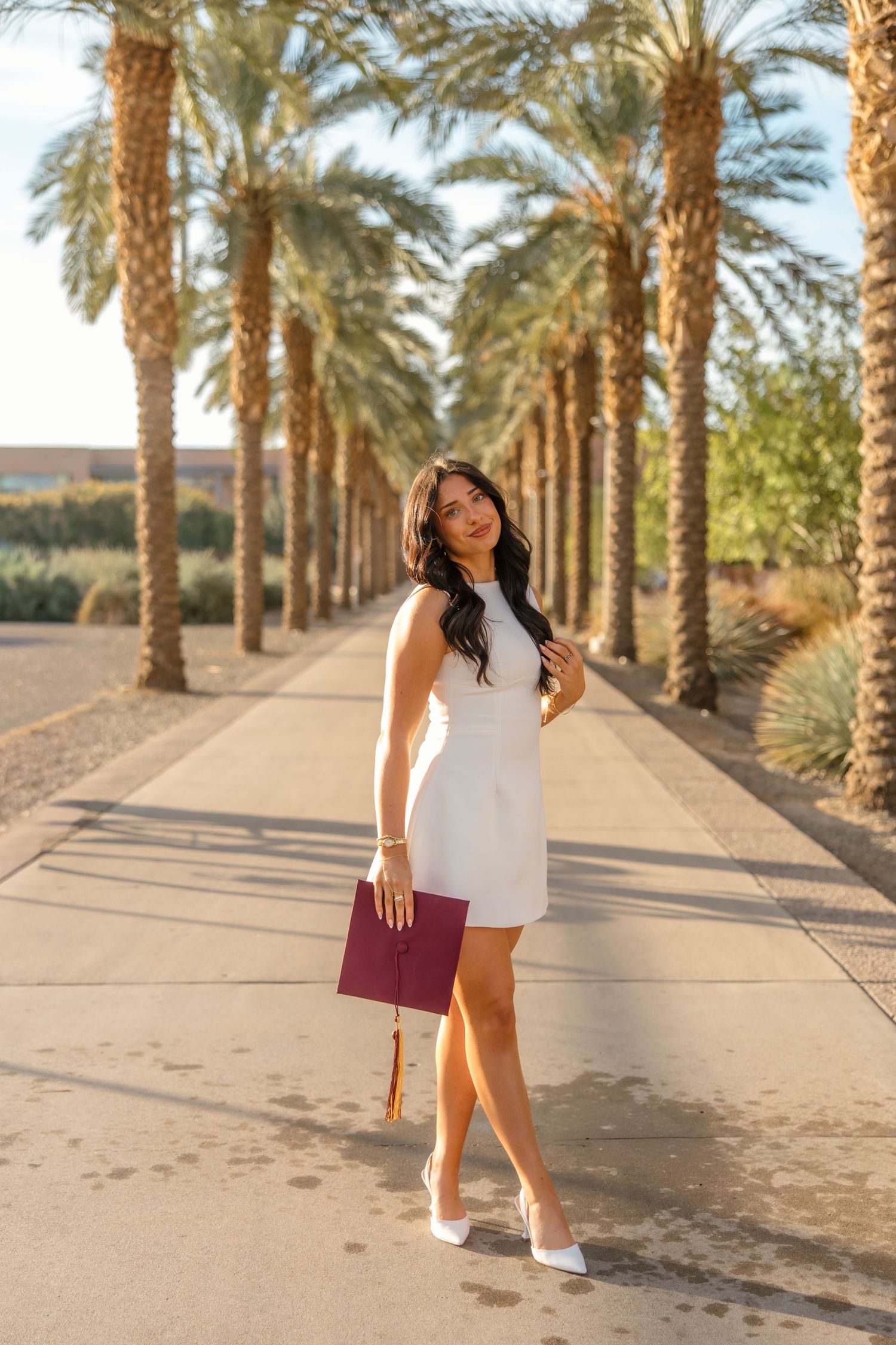 Woman in white dress holding graduation cap walks palm tree-lined path in golden hour light.