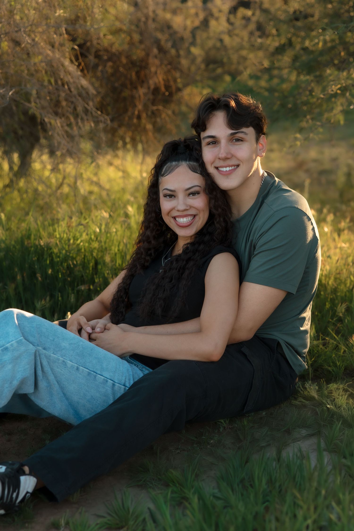 Smiling young couple sitting together on grass in warm sunlight with arms around each other.