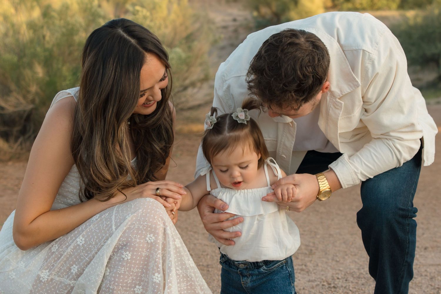 Young family with parents in white outfits crouching down to interact with their toddler girl in desert setting.