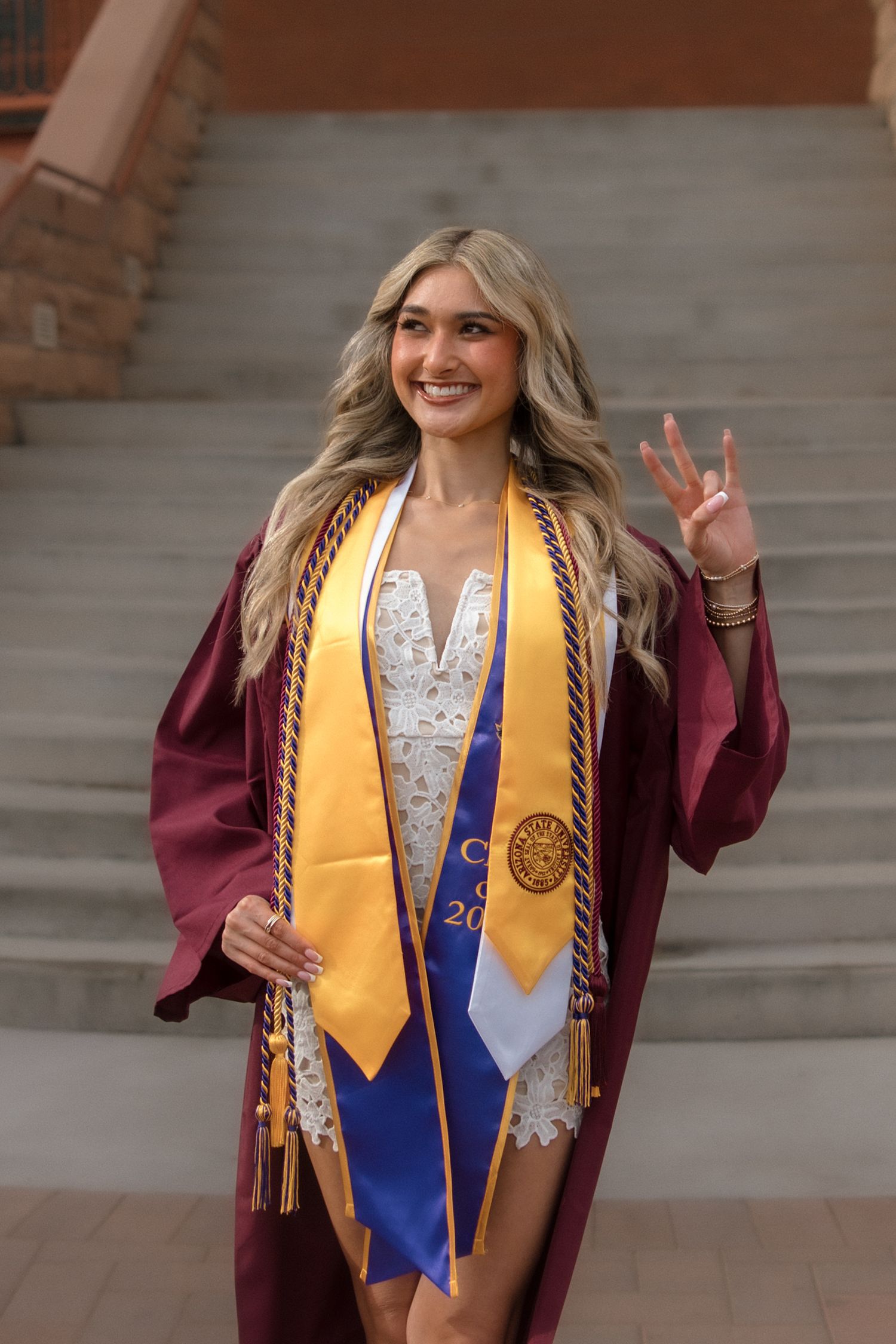 Blonde ASU graduate in maroon graduation robe and gold honor stole making pitchfork hand sign on campus staircase.