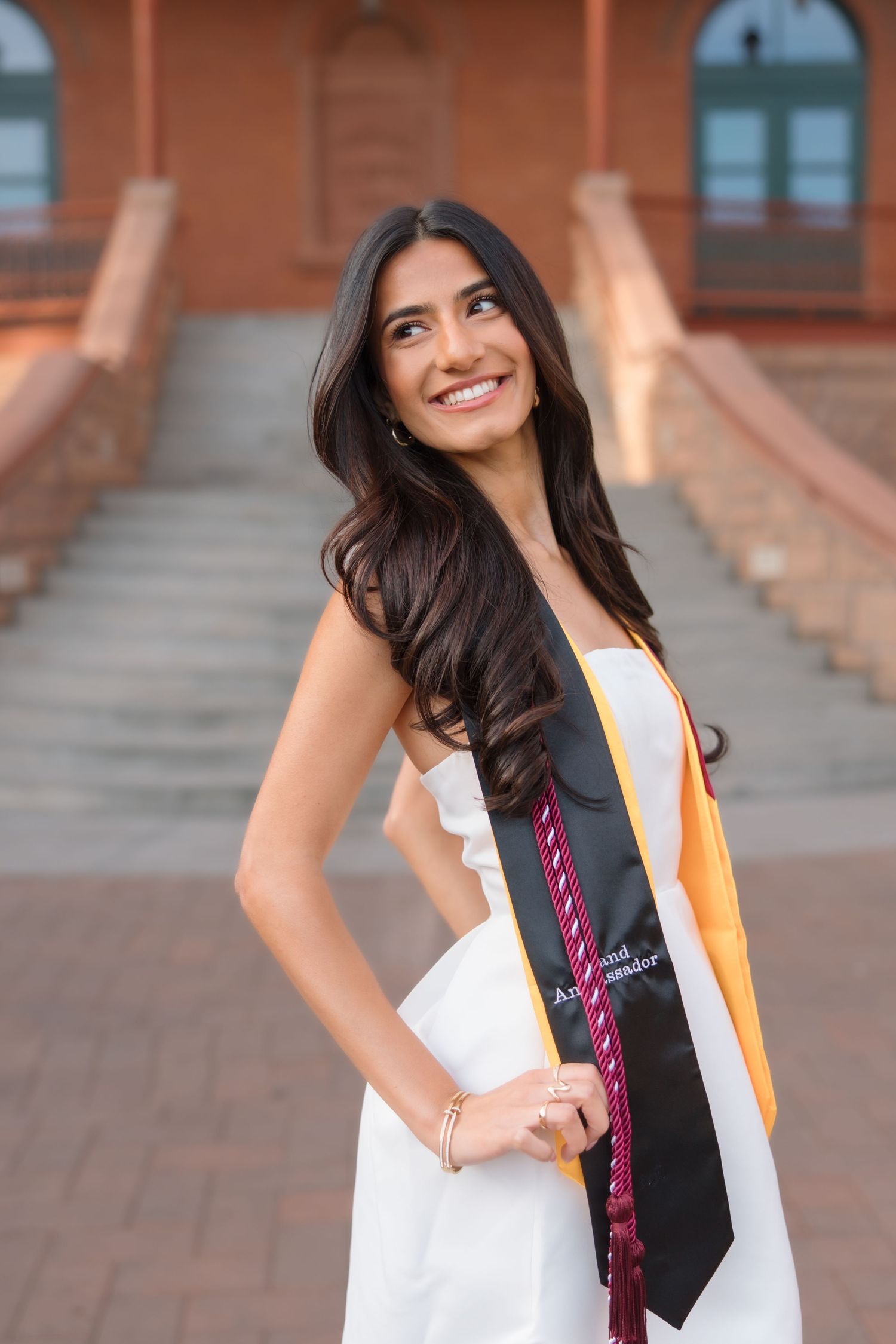 Smiling female graduate in white strapless dress with black and gold honor stole on campus steps.