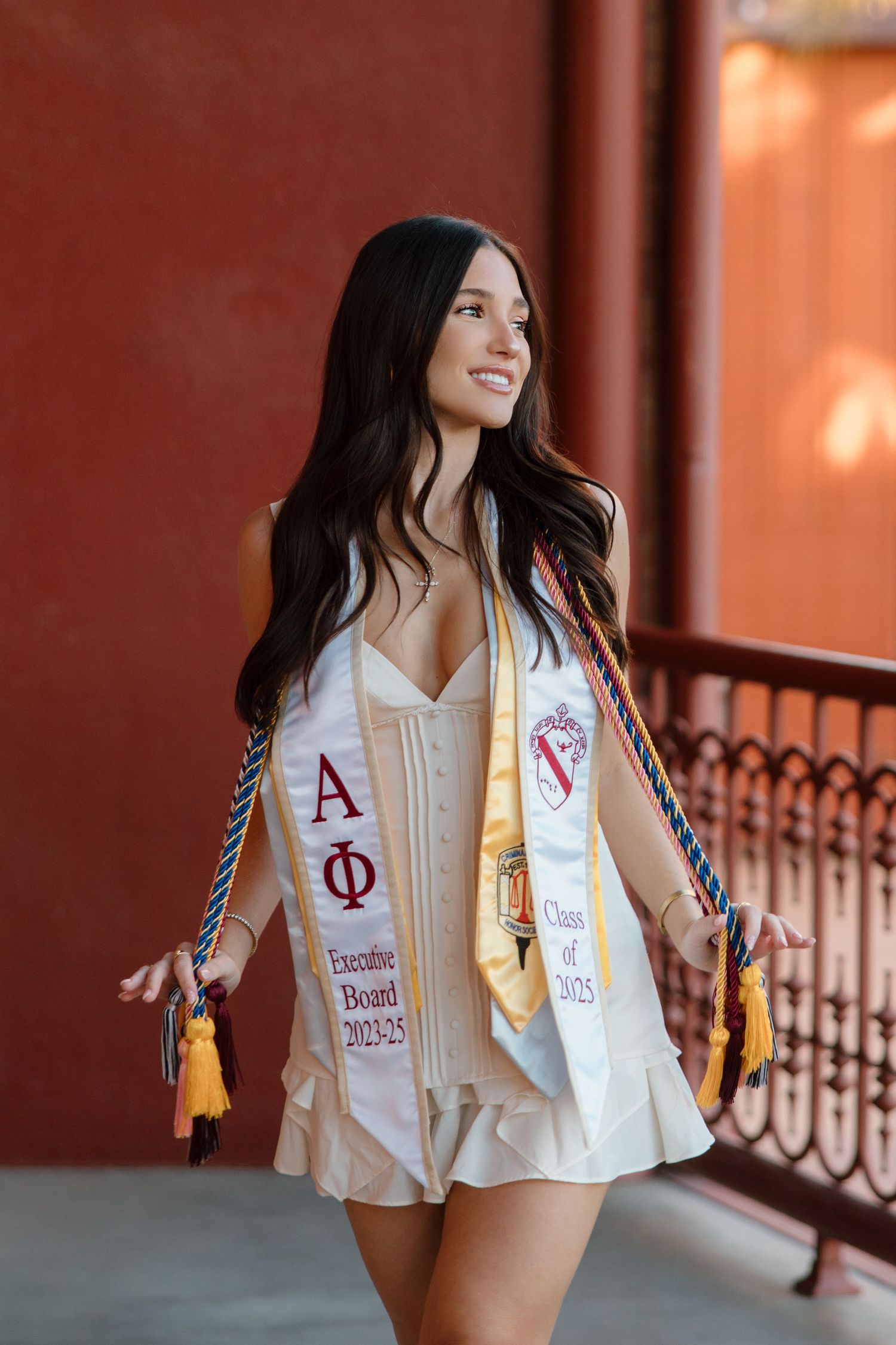 Smiling female graduate wearing Alpha Phi stole and Class of 2025 sash walks on red brick campus building.