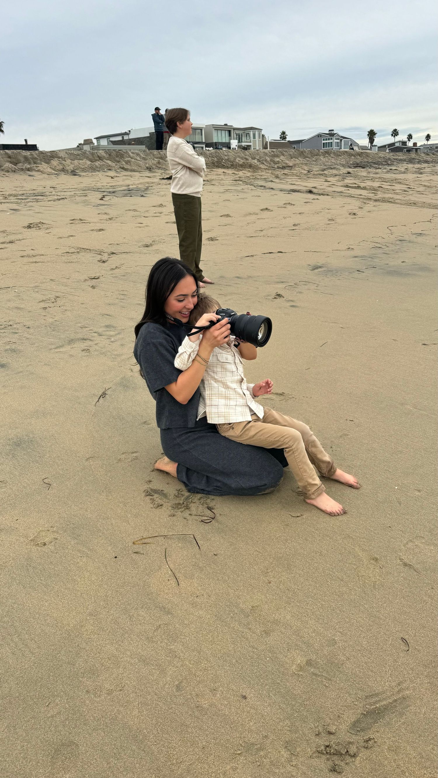 Woman photographer sits on beach sand with toddler looking through camera lens on overcast day.