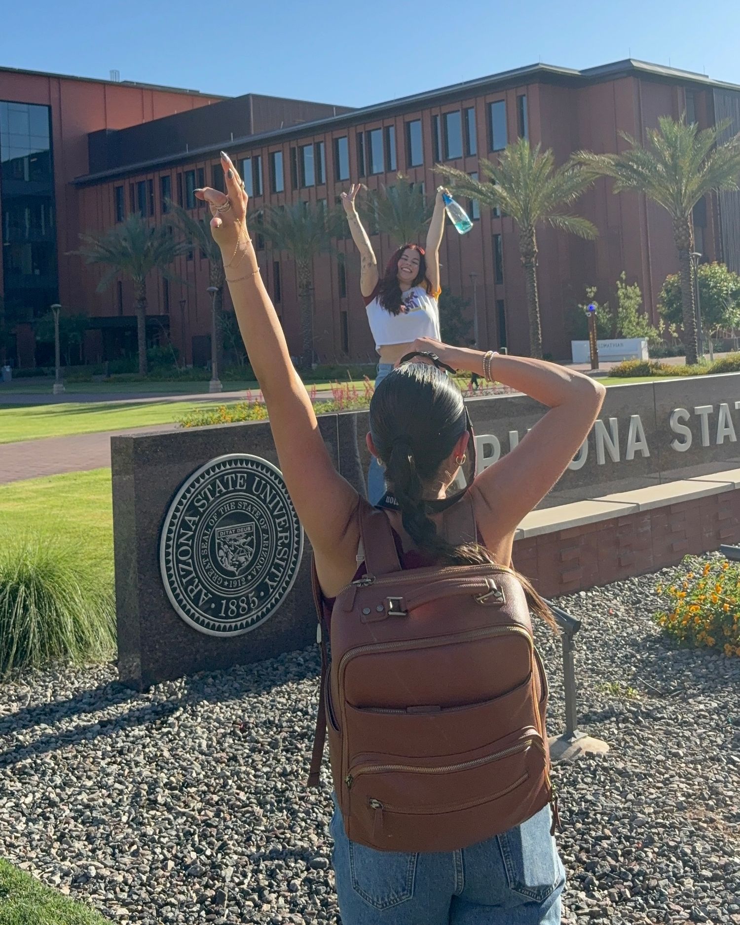 Photographer gestures to student posing at Arizona State University entrance sign on sunny day.