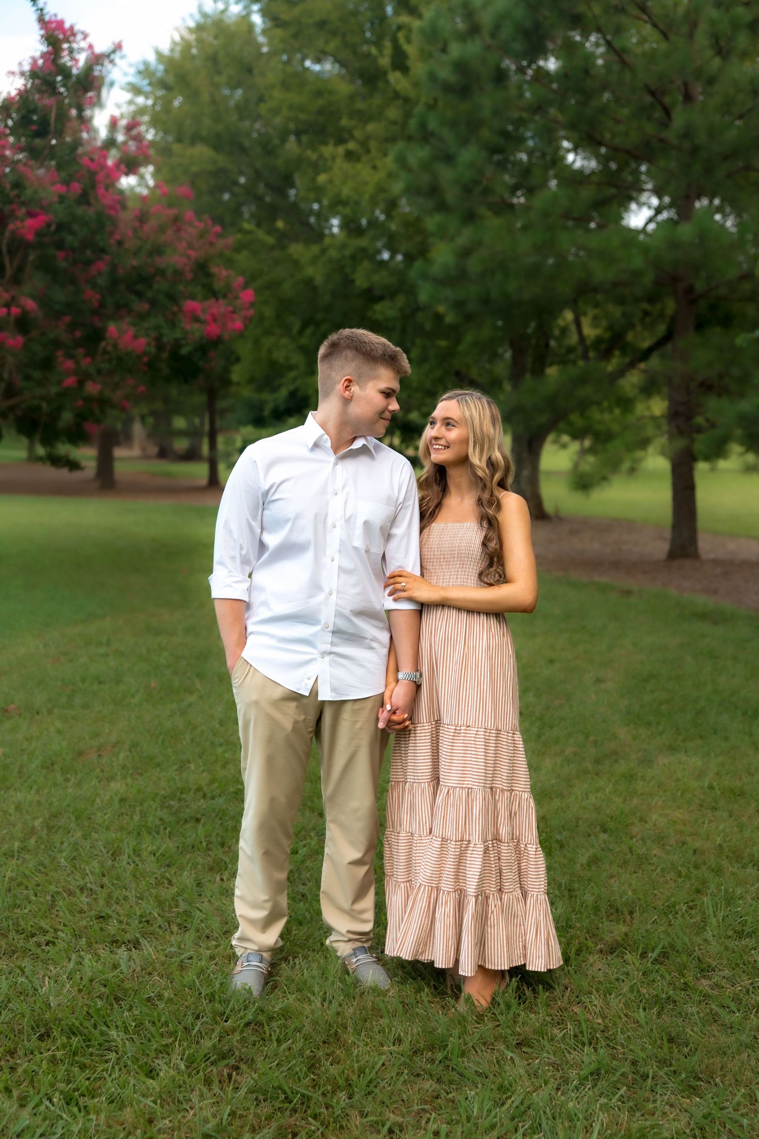 Couple holding hands smiling at each other in a lush green park, man in white shirt and woman in pink maxi dress.