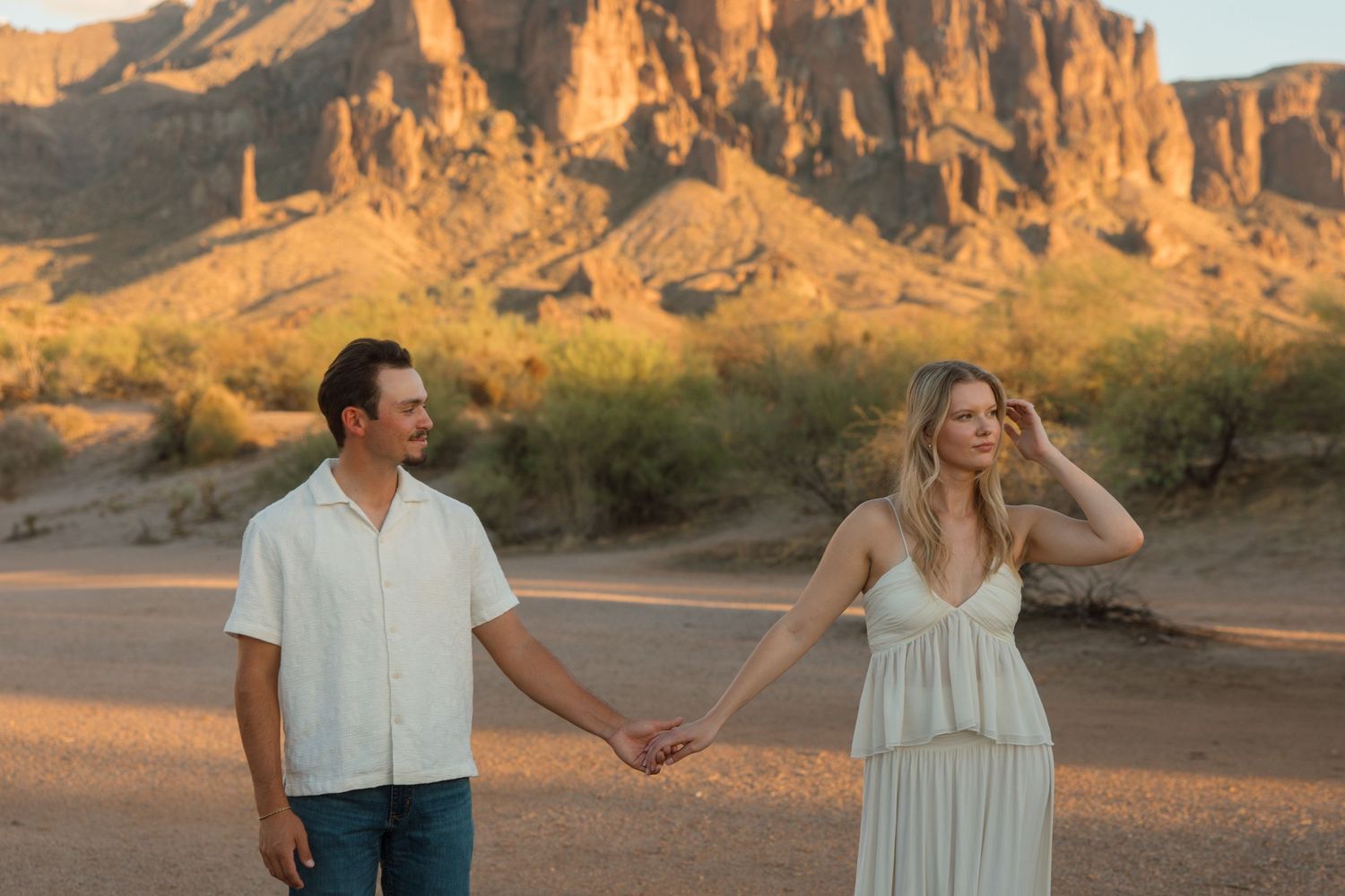 Couple holding hands in desert landscape with dramatic rocky mountains at golden sunset hour.