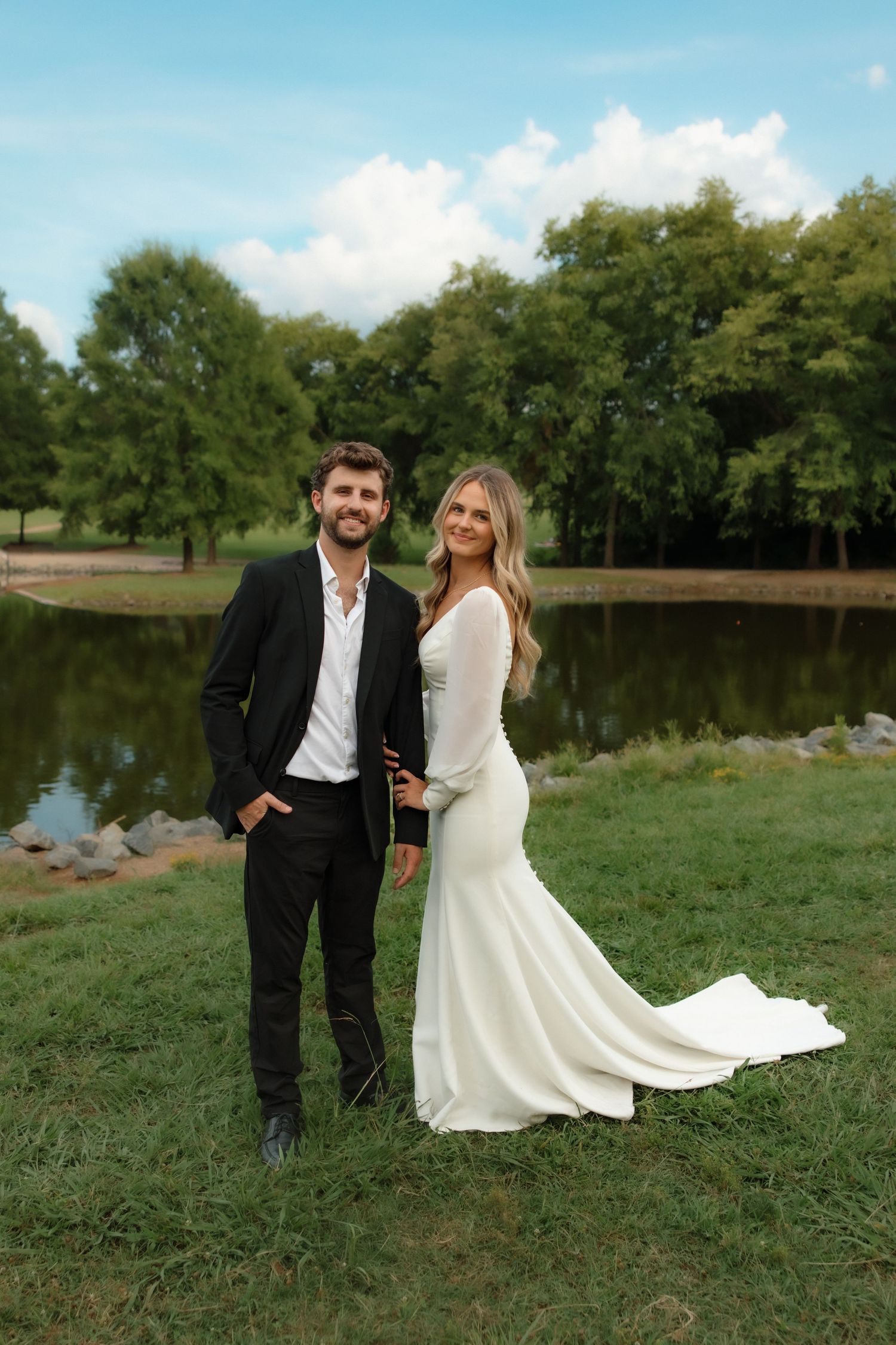 Bride and groom smiling together by a serene lake, she in a white gown, he in a black suit.
