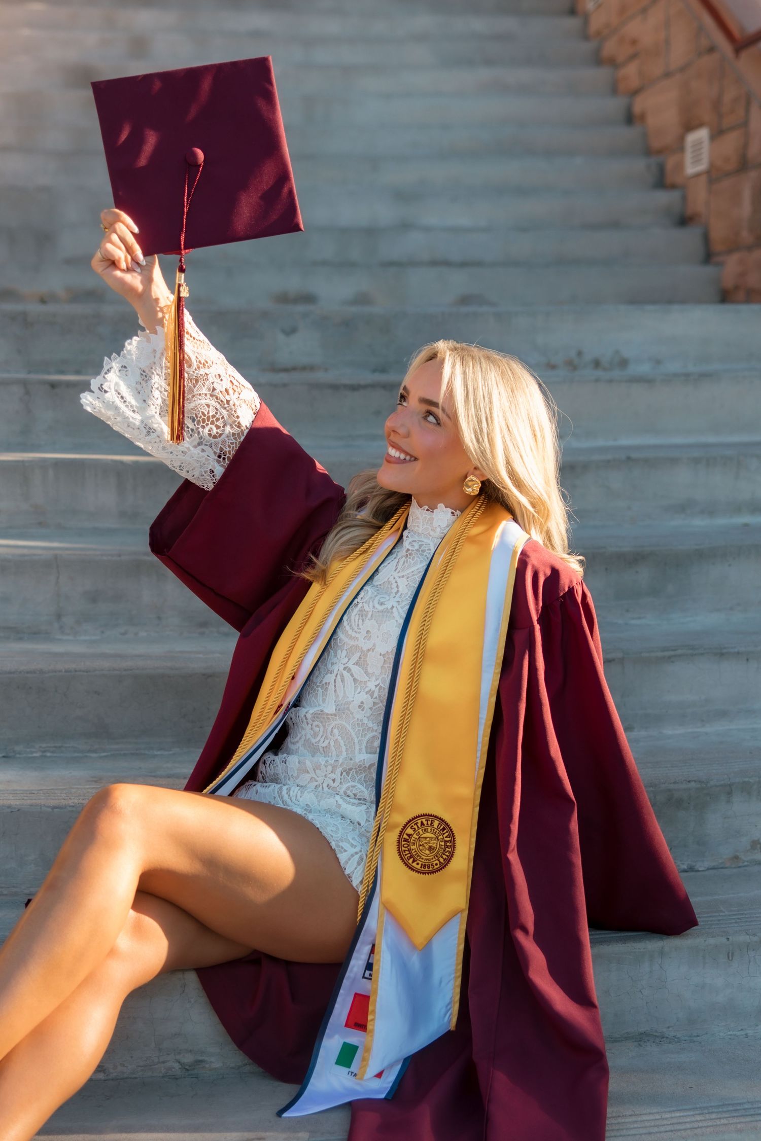 Smiling blonde graduate sitting on stone steps holding up maroon cap and gown with gold honor stole.