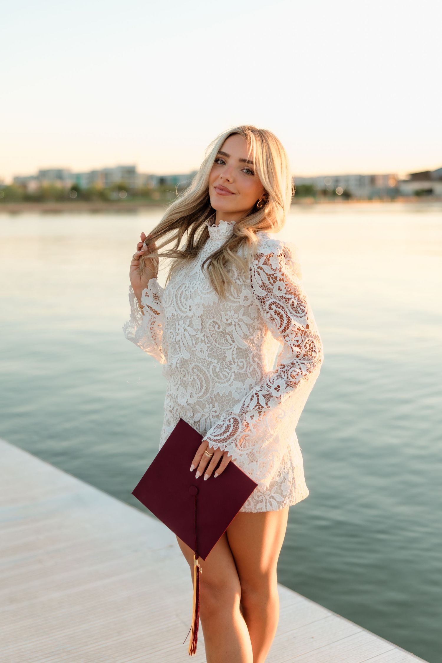 Blonde woman in white lace mini dress holding maroon clutch posing on waterfront dock at sunset.