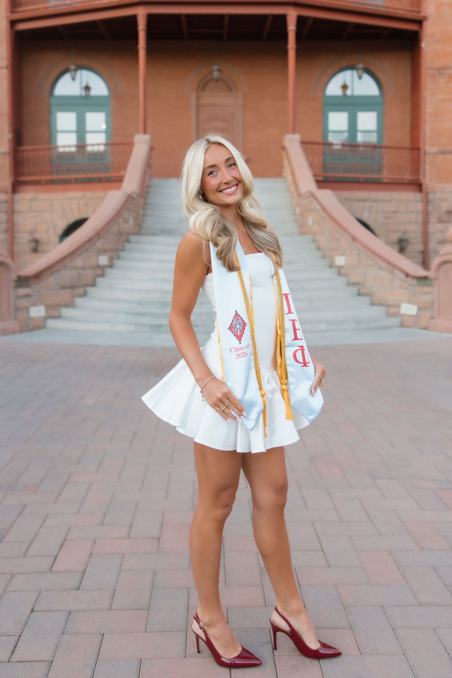 Smiling blonde graduate in white dress with Pi Beta Phi sash and gold honor cord poses outdoors.