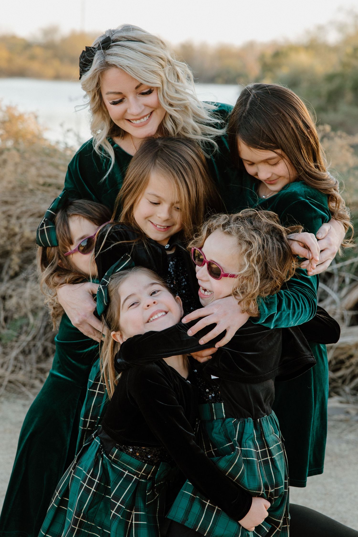 A smiling blonde woman in green velvet hugs four young girls in matching holiday outfits outdoors.