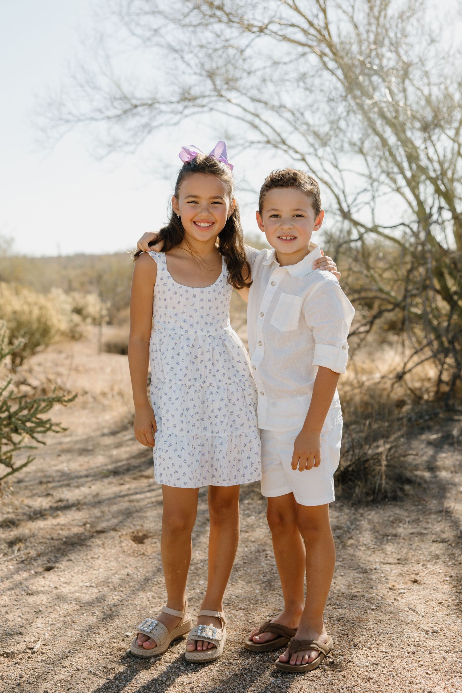 Two smiling children in white outfits wearing Easter bunny ears pose together on a sunny desert trail.