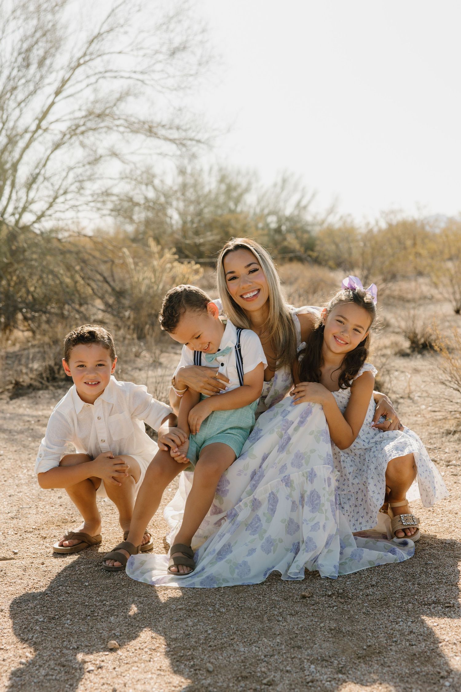 A smiling woman sits outdoors in a desert setting with three young children, all dressed in light pastel outfits.