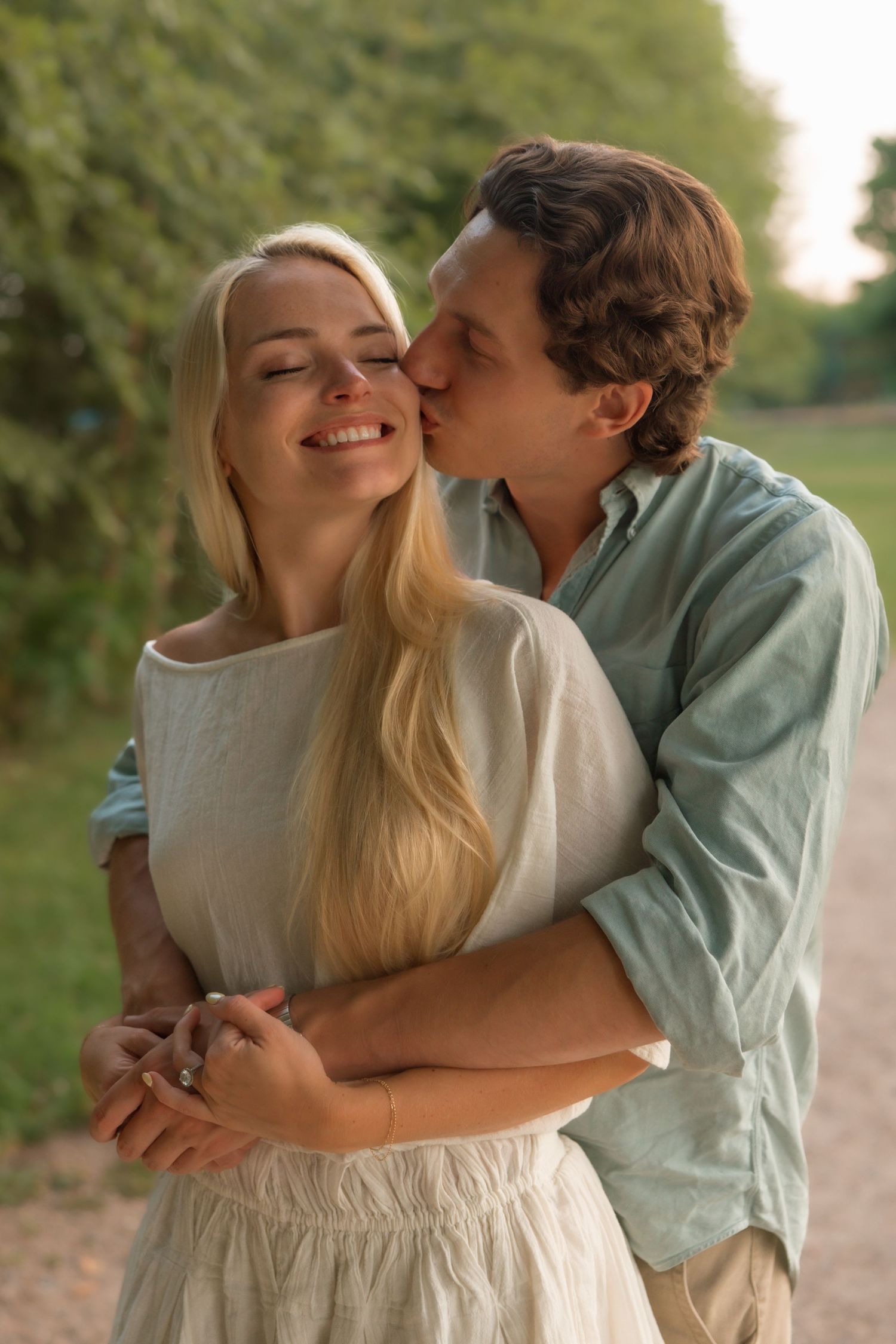 Man in blue shirt kissing woman's cheek while embracing her from behind on a sunny outdoor path.