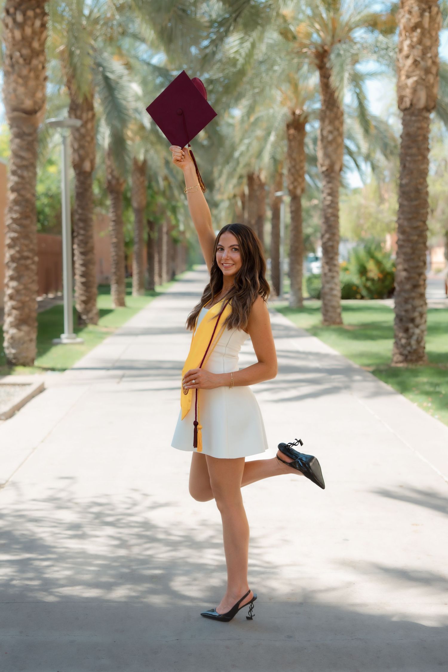 Joyful female graduate in white dress raising maroon cap along palm tree lined walkway at graduation.