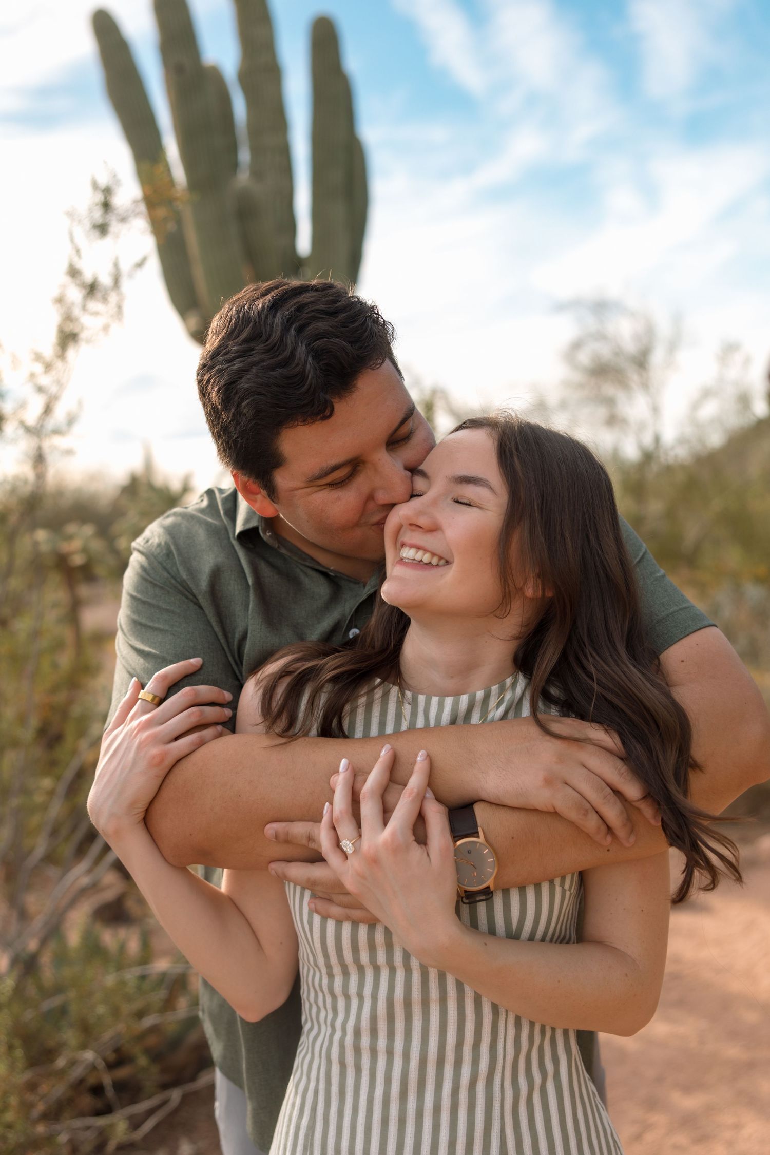 Man hugging woman from behind and kissing her cheek while she smiles near a saguaro cactus.