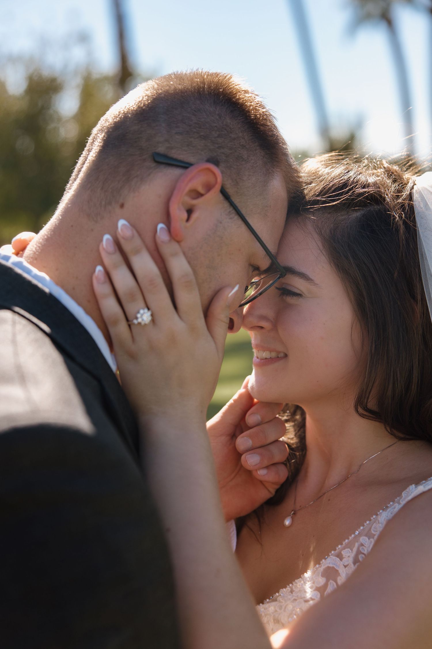 Bride and groom share a tender forehead touch outdoors with palm trees behind them.