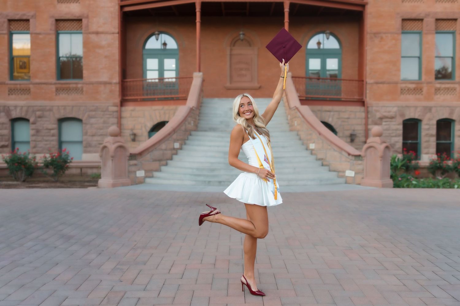 Young woman in white dress tossing graduation cap in front of brick university building steps.