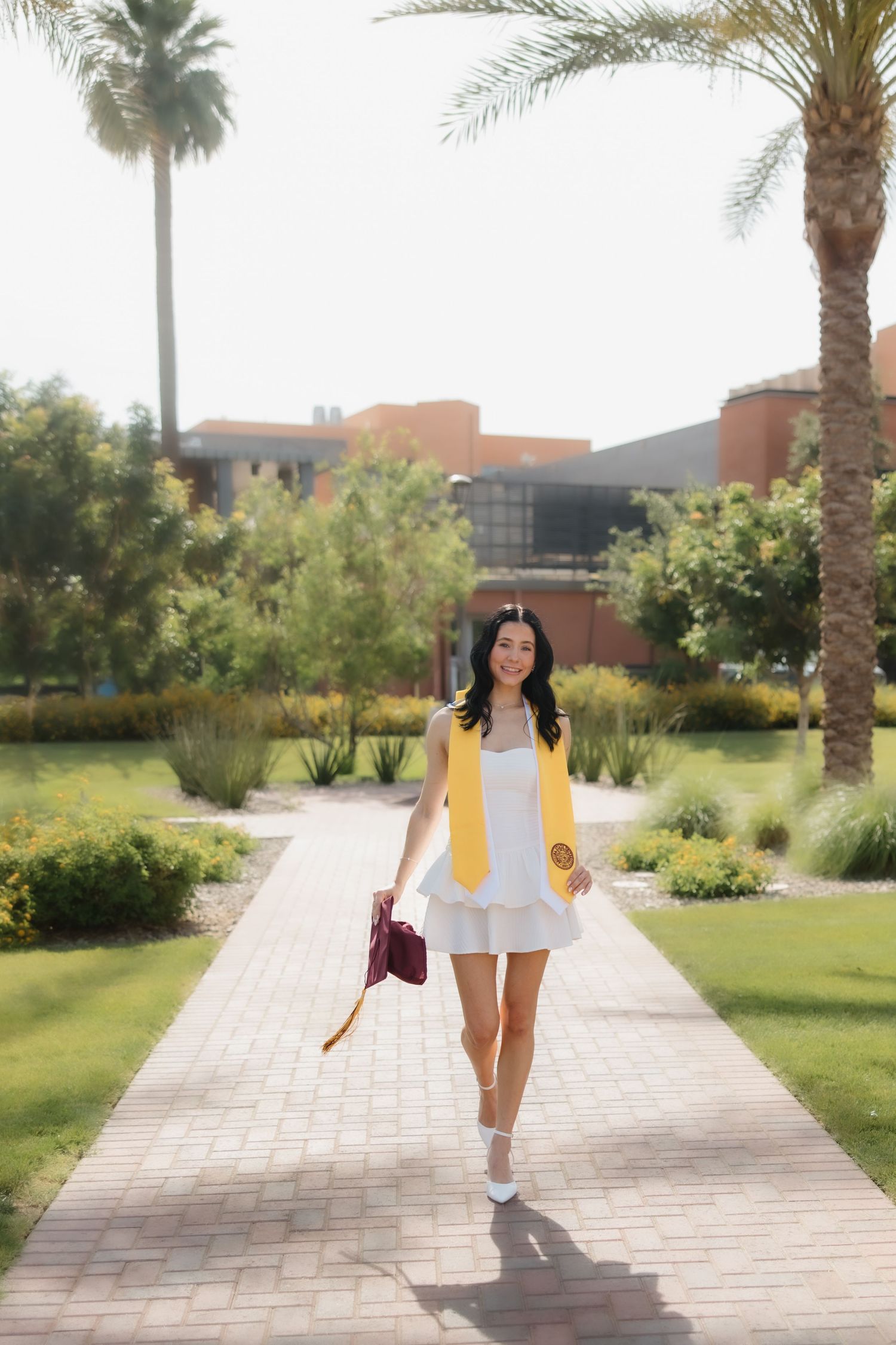 A young woman in a white dress and yellow graduation stole walks along a palm-lined campus pathway.