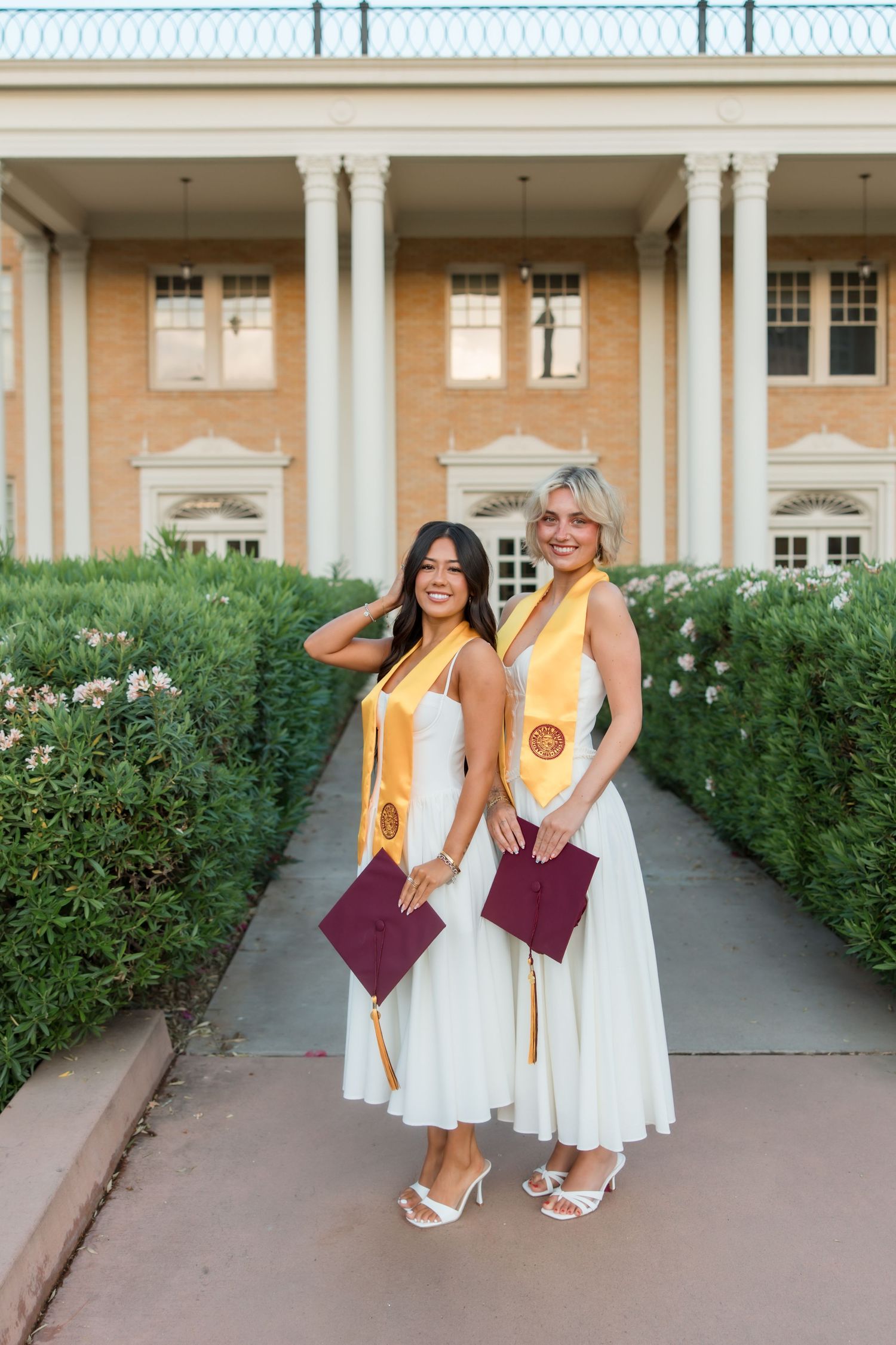Two smiling graduates in white dresses and gold stoles hold maroon caps outside a brick building.