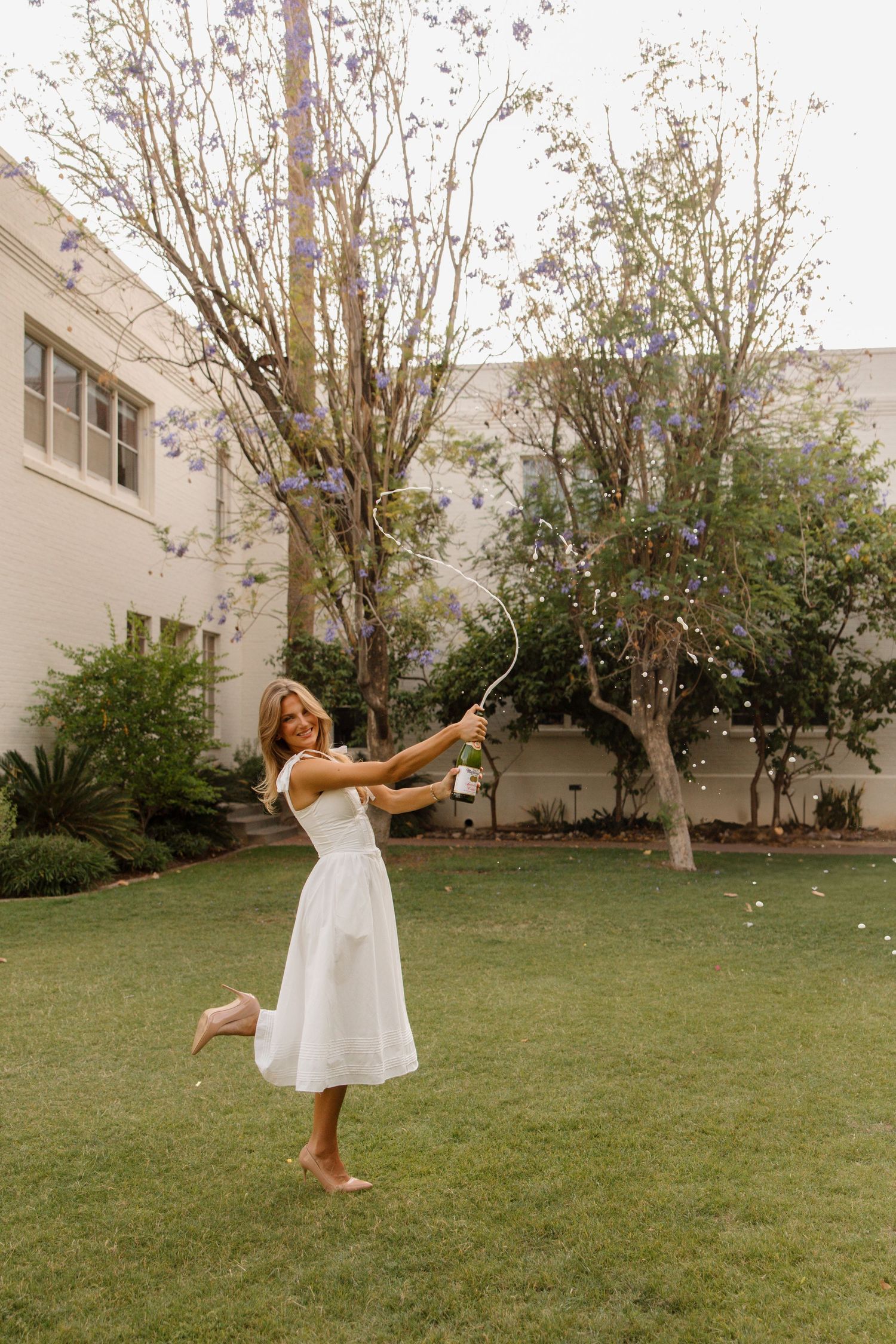 Woman in white midi dress twirling barefoot on green lawn, popping champagne confetti cannon near flowering trees.
