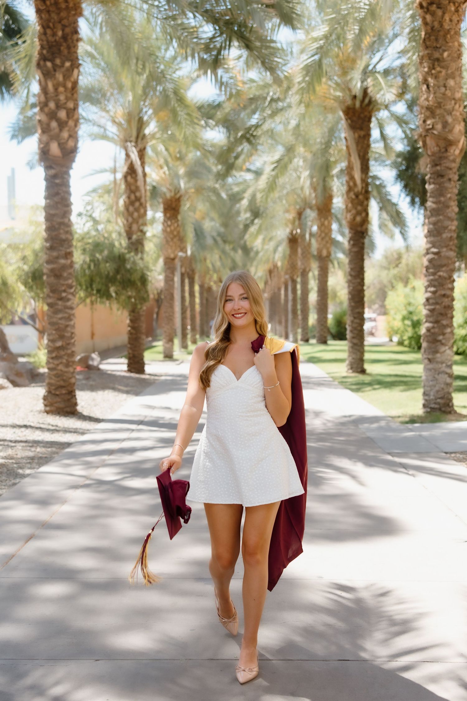 A smiling graduate in a white romper holds a maroon cap and gown while walking among palm trees on a sunny day.