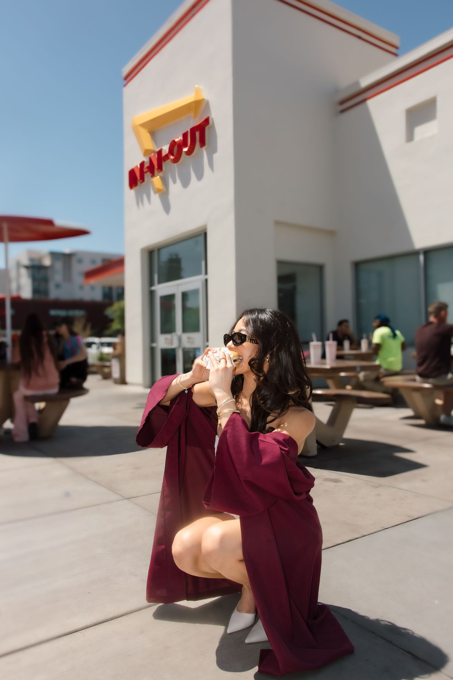 Graduate in maroon gown crouching and eating a burger outside In-N-Out Burger restaurant.