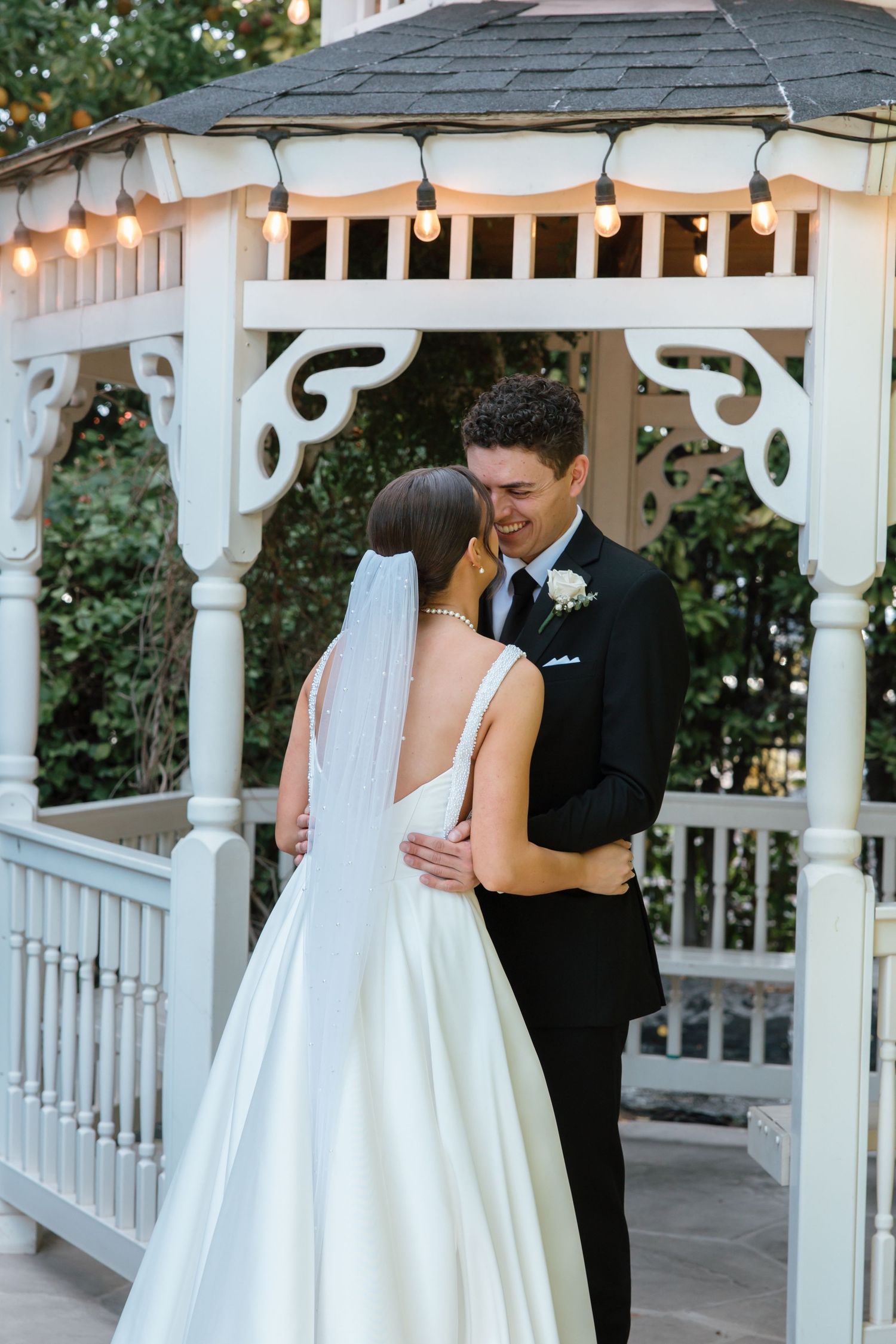 Smiling groom embraces bride in white gown with veil under a white gazebo with string lights.