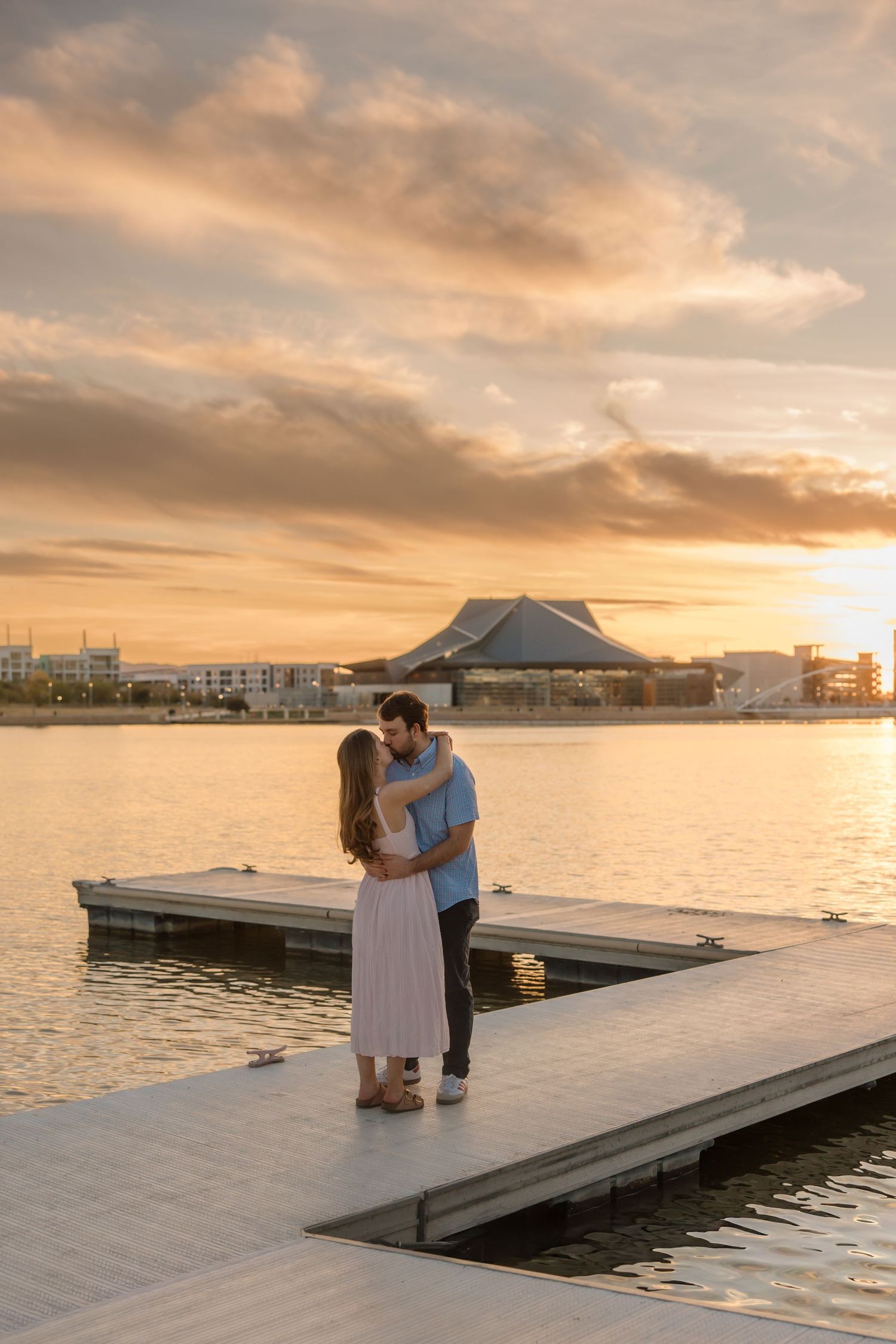 Happy couple hugs on wooden pier at golden sunset with scenic waterfront building reflecting on calm water.