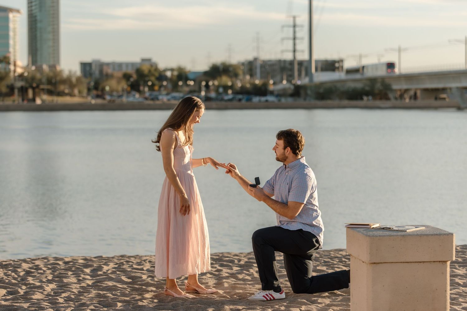 Man proposing to woman on one knee at a scenic waterfront beach during golden hour with city skyline backdrop.