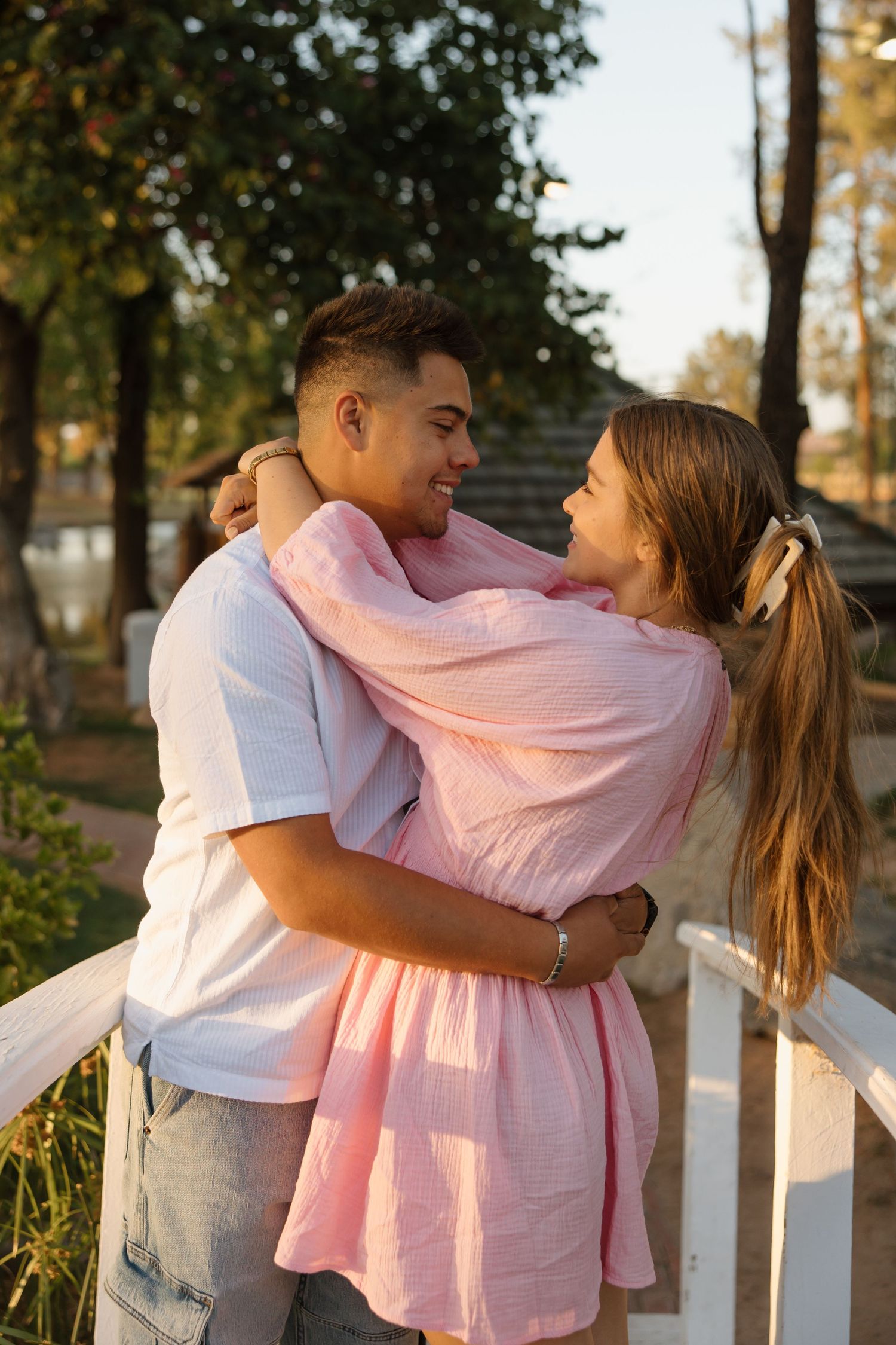 A young couple embracing and smiling at each other outdoors near a white fence, surrounded by trees in warm sunlight.