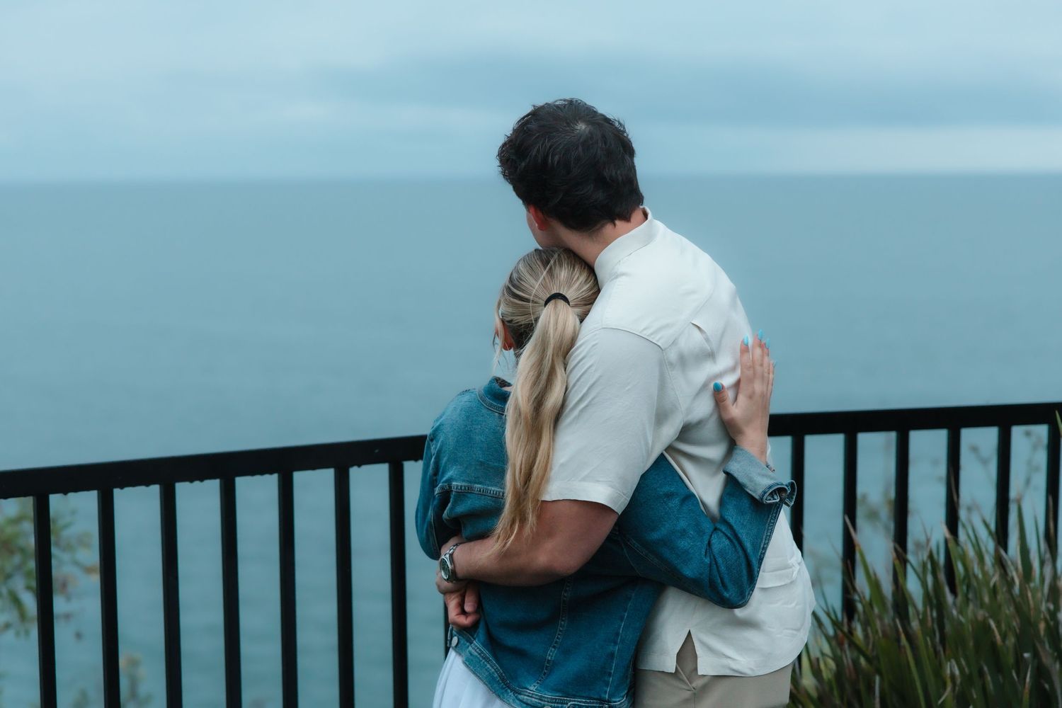 Engaged couple embracing while gazing at the ocean from a scenic overlook together.