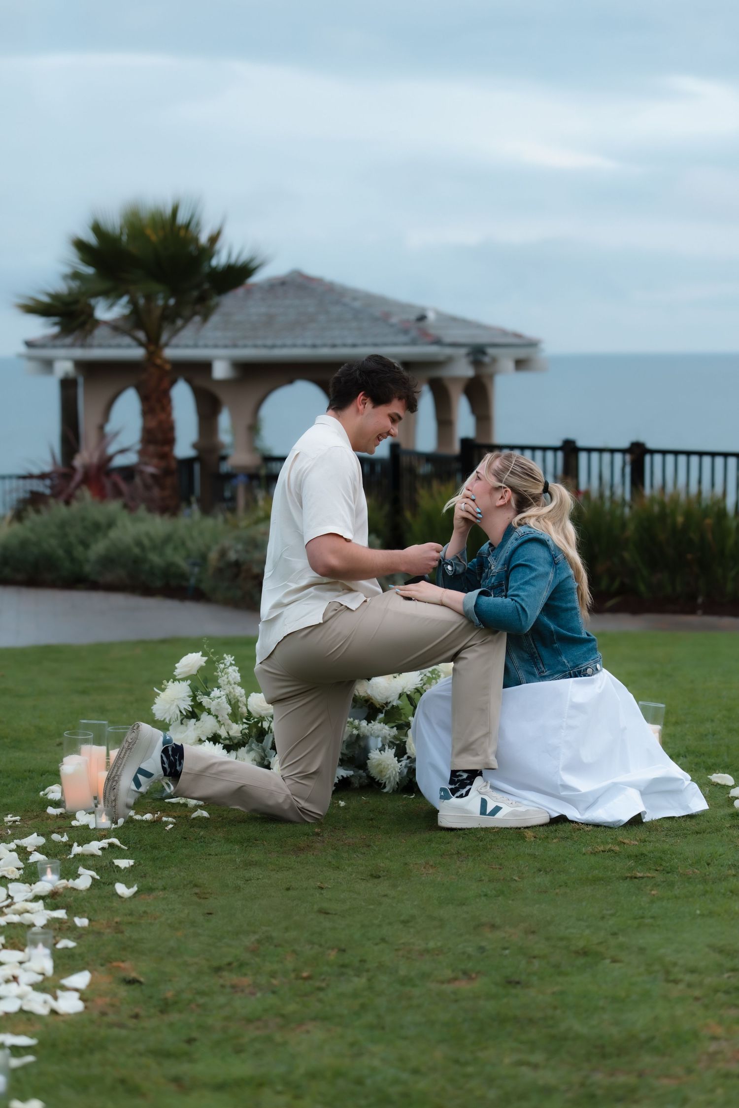 Romantic marriage proposal on lawn with rose petals, candles, and white flowers by the sea.