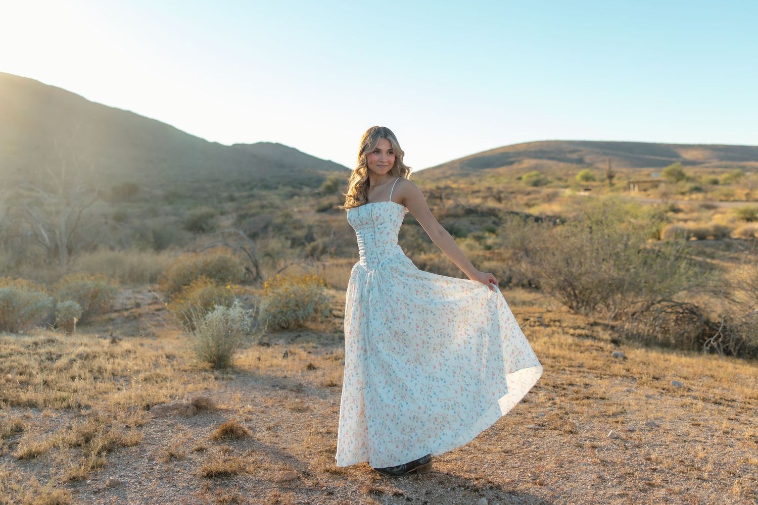 Blonde woman in white floral maxi dress posing in desert landscape at sunset.